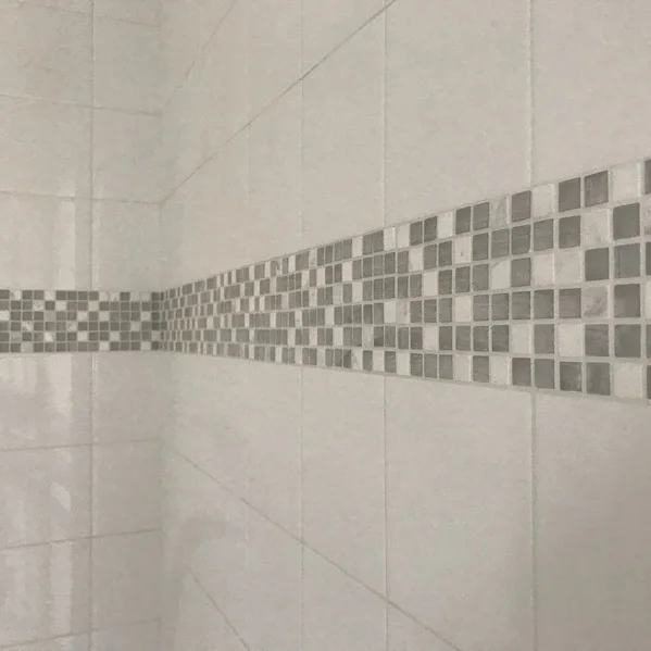 Close-up of a tiled wall with a horizontal strip of small, square mosaic tiles in various shades of gray.