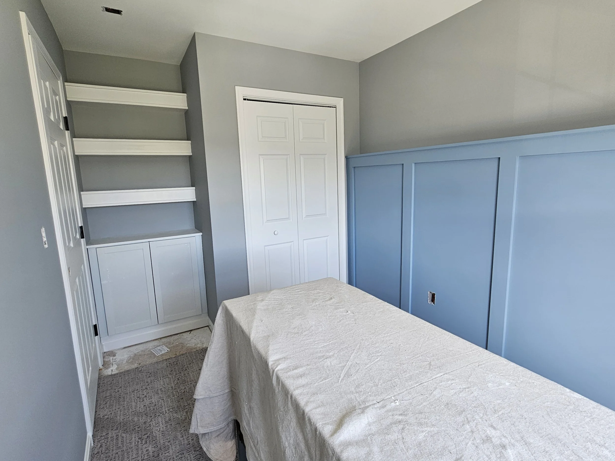 An empty room with gray walls, a white built-in cabinet with shelves, a closed white closet door, and a blue wainscoting on one wall. There is a table covered with a light-colored cloth in the foreground.