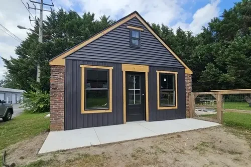Small modern house with black siding, yellow trim around the windows and door, brick sides, and a concrete sidewalk in front, with trees and a blue sky in the background.