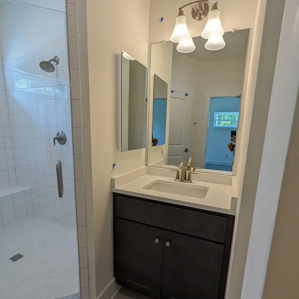 Bathroom with a shower area on the left, a vanity with a sink and dark cabinet underneath, a large mirror, and a three-light fixture above. A reflection in the mirror shows a door and a window in the background.