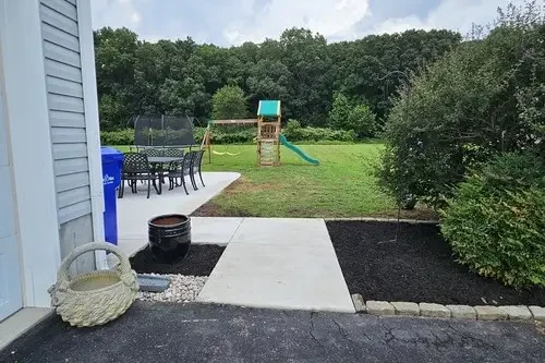 Backyard with a concrete path, outdoor furniture, a child's playset, and a lawn surrounded by bushes and trees.