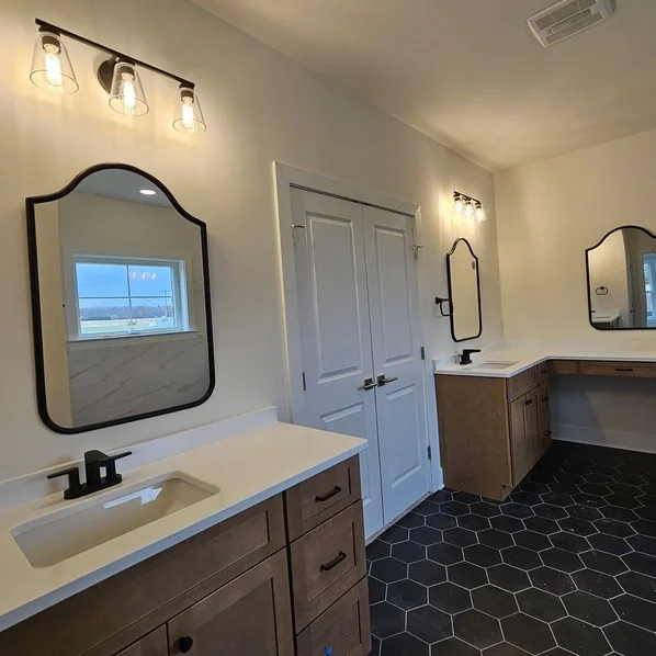 Bathroom with two white vanities, each with a black faucet and a rectangular mirror, black hexagon tile floor, white walls, and a window showing a blue sky outside.