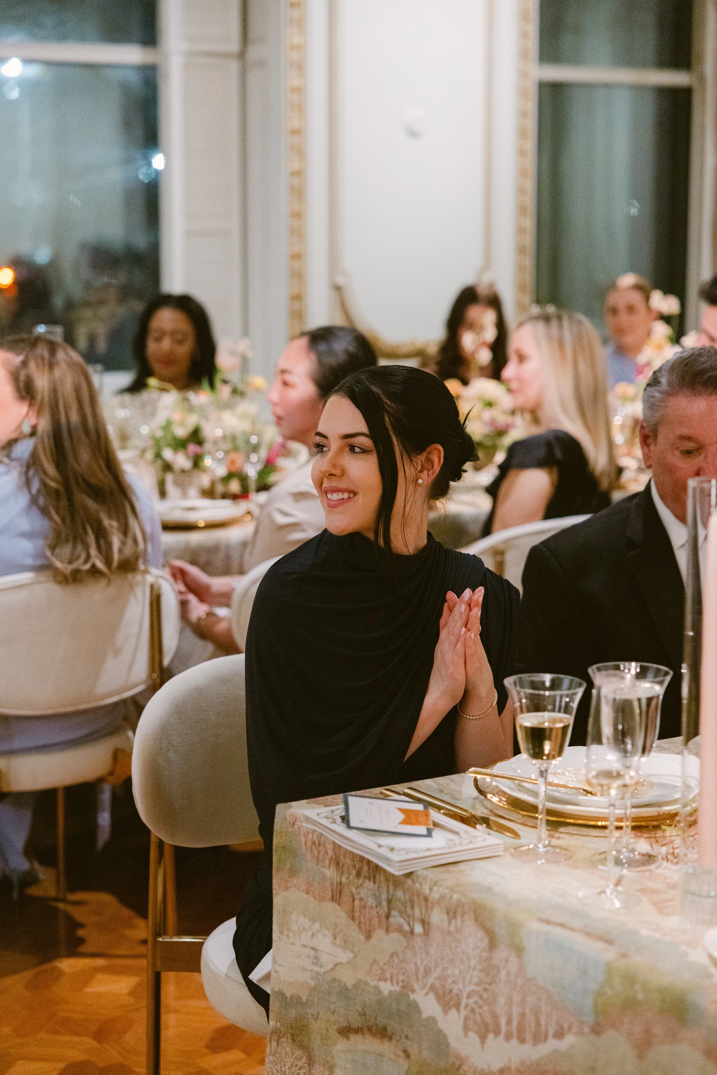 A woman wearing a black dress sitting at a round table, clapping her hands, with a smile on her face. The table has a glass of champagne and some papers or a menu. The setting is a formal dinner with several other people in the background, decorated with floral arrangements.
