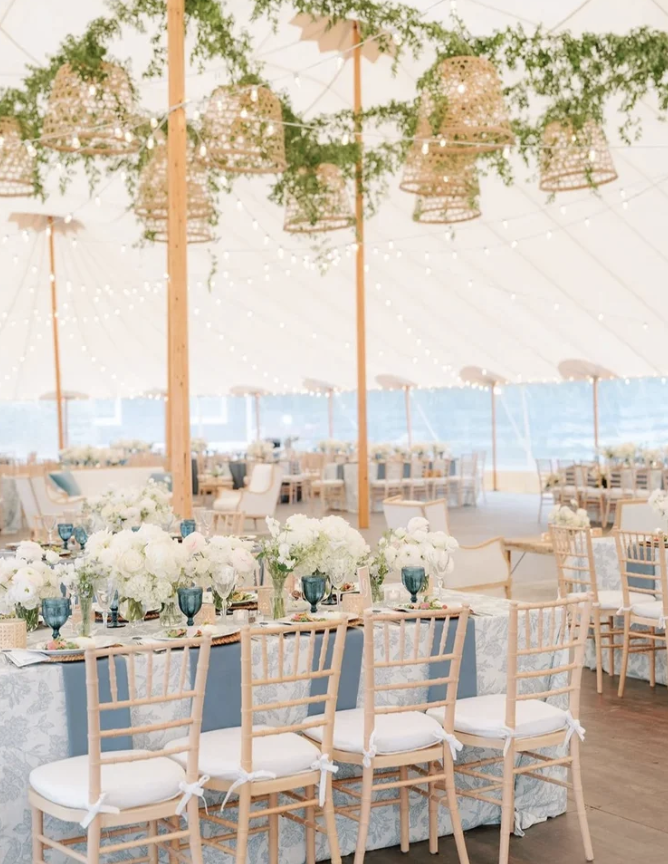 A beautifully decorated outdoor event tent with long tables covered with floral centerpieces and blue glassware. Wicker lanterns and string lights hang from the ceiling, creating a warm ambiance. Several chairs are arranged around the tables, ready for guests.