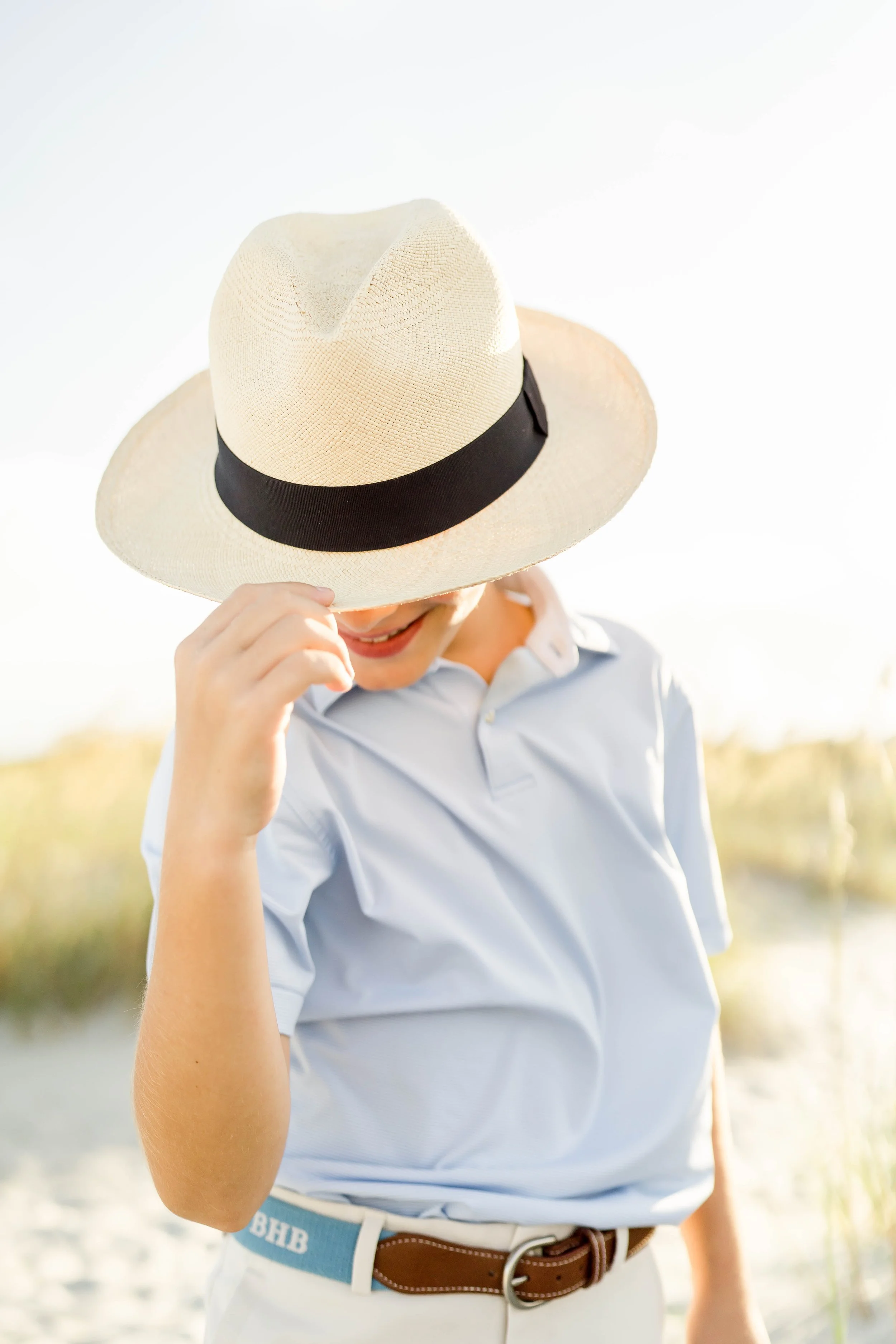 A person wearing a white hat with a black band, a light blue polo shirt, and a brown belt, standing outdoors in a bright sunny setting.