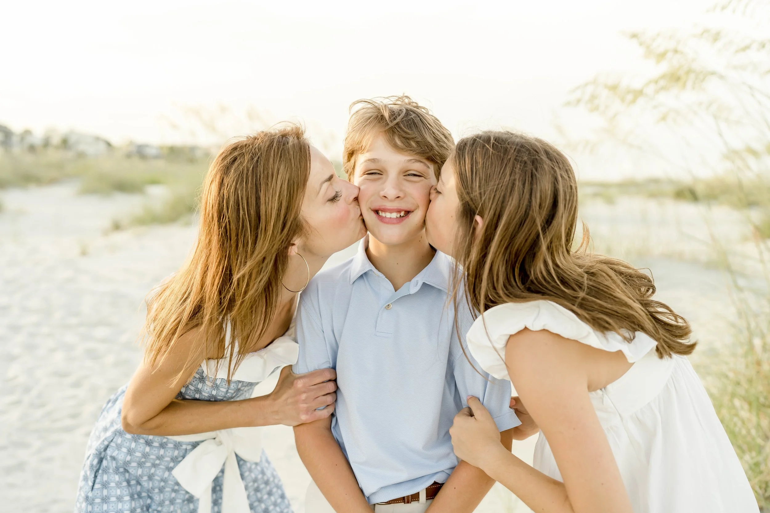 Three young people, two women and a boy, sharing a kiss and hug on a beach with a blurred natural background.