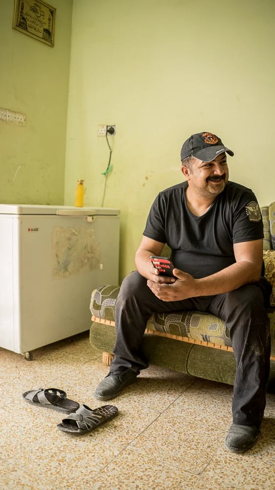 A man sitting on a small couch in a room with pale green walls, holding a mobile phone, smiling and looking to his right. He is wearing a black T-shirt, black pants, black shoes, and a black baseball cap with orange logo. On the floor in front of him are a pair of black flip-flops and a remote control. There is a small white refrigerator with a yellow bottle on top and a wall socket with a green plug in the background.