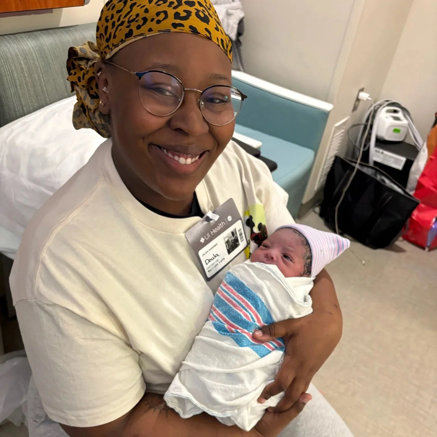 A smiling woman wearing glasses and a leopard print headscarf holding a newborn baby wrapped in a hospital blanket.