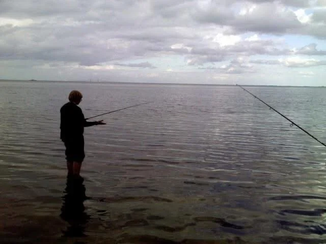 A person standing in shallow water by a lake or ocean, fishing with a rod, under a cloudy sky.
