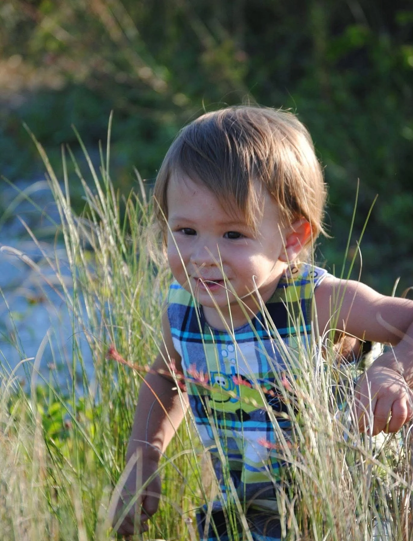 A young boy with light brown hair and dark eyes crouches in tall grass near water, wearing a colorful plaid shirt.