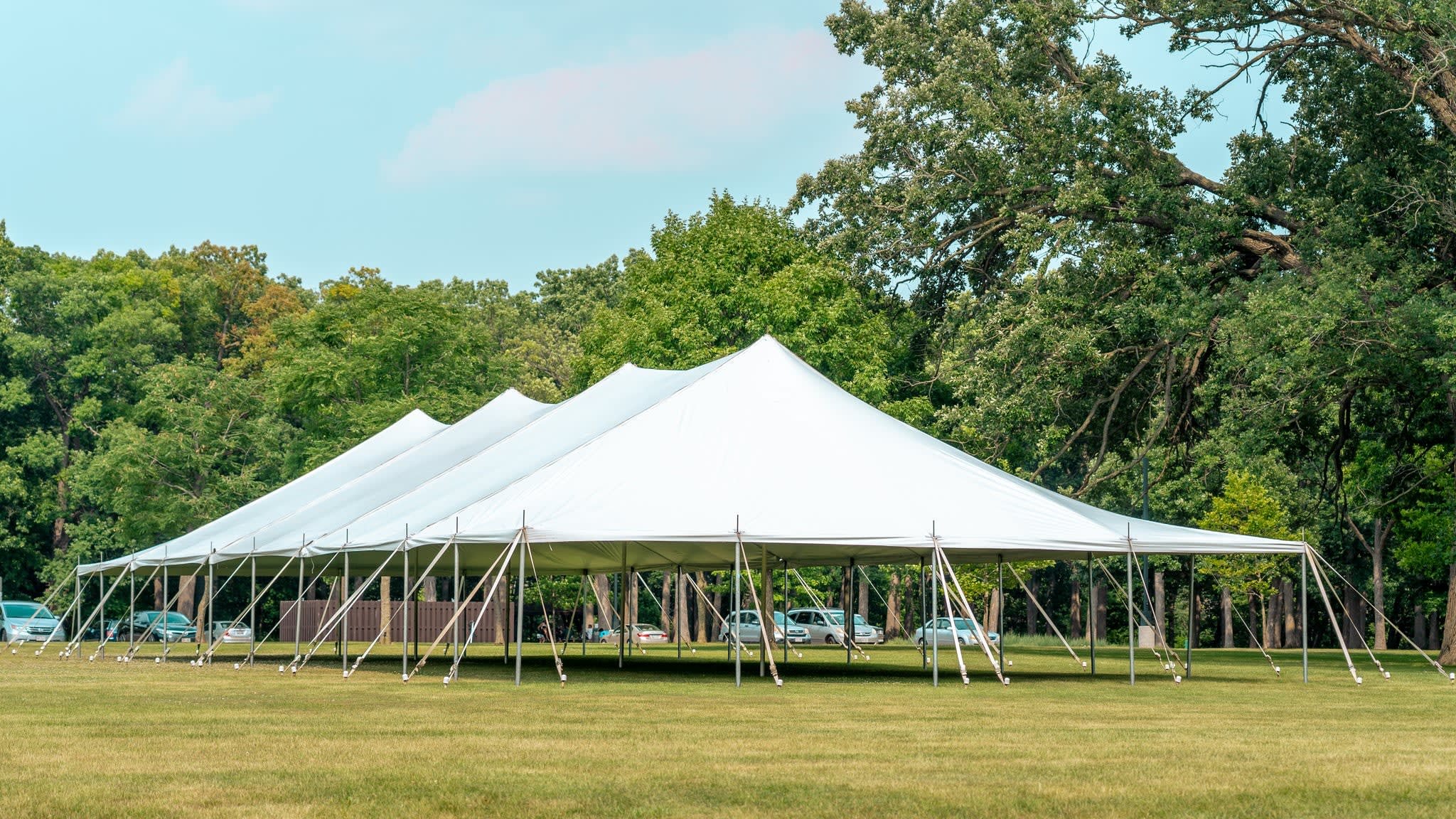 Large white event tent set up on grassy area with rows of contingency parking lot with several cars and lush green trees in the background.