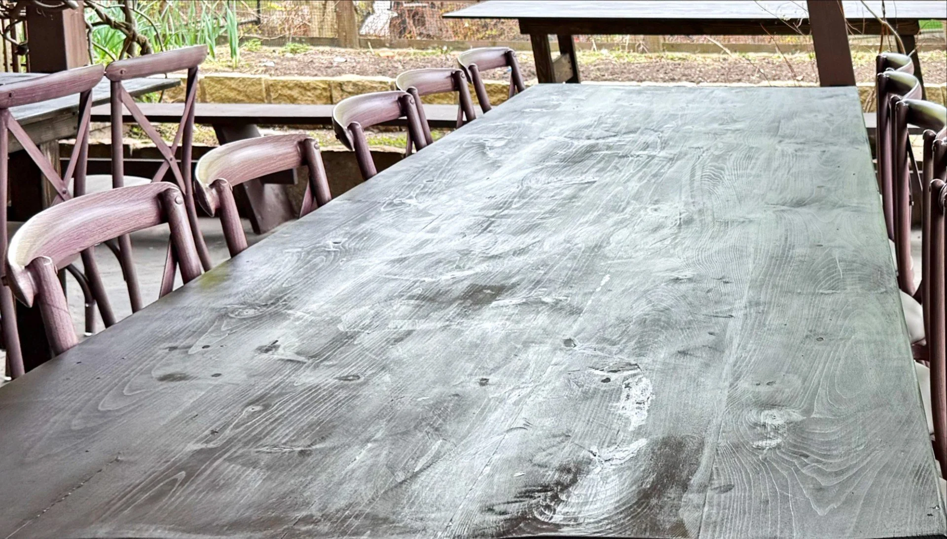 Unfinished wooden table with visible wood grain in an outdoor patio area, surrounded by wooden chairs with brown cushions, with a garden and stone wall in the background.