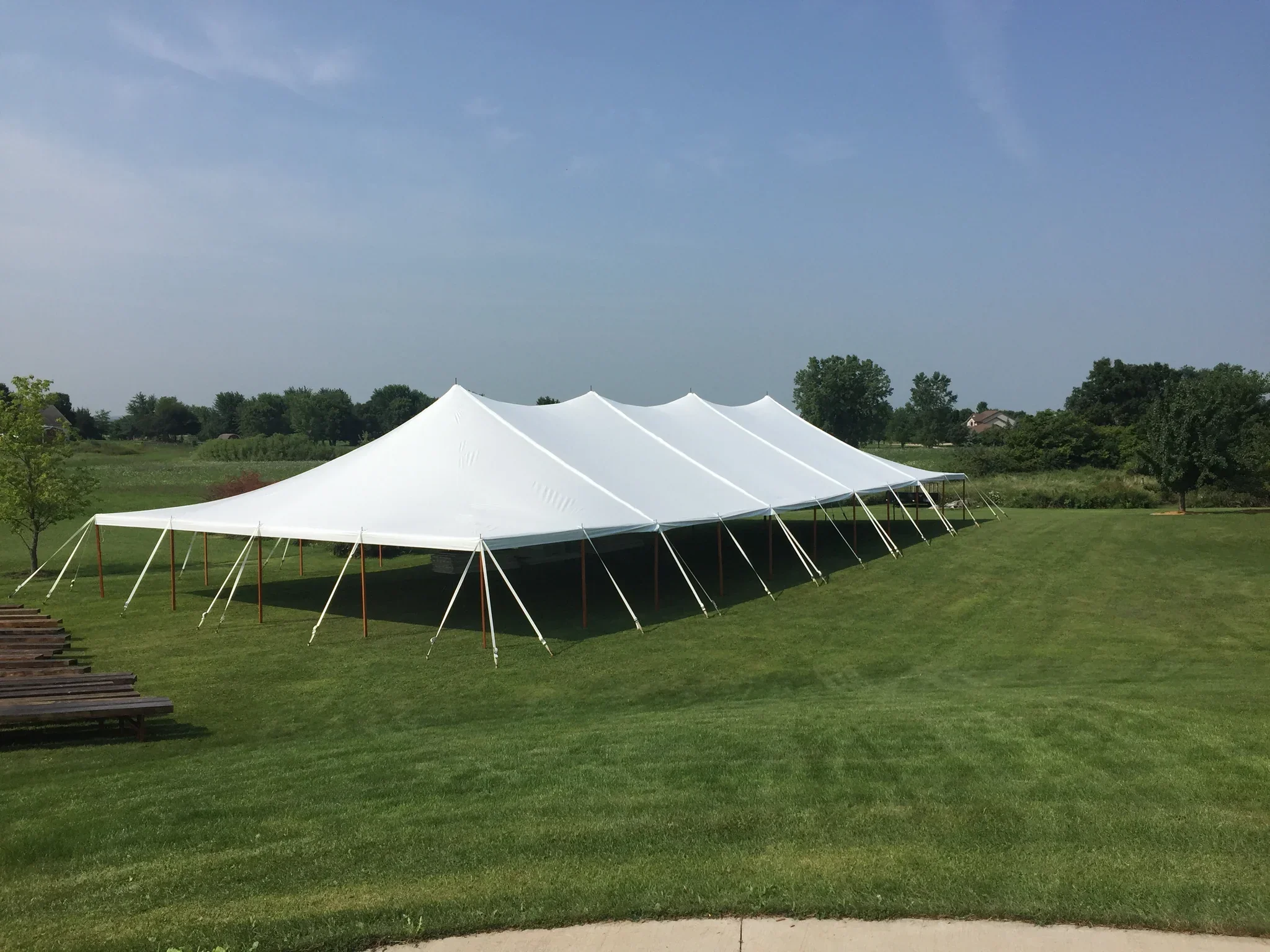 A large white event tent set up on green grass in a park or open outdoor area, with trees and houses in the background under a blue sky with some clouds.