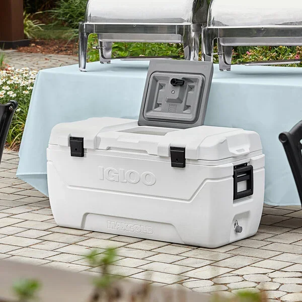 A white portable cooler with a lid open, placed on a paved outdoor patio. The cooler has black latch closures and a handle on the side. It is positioned in front of a table with a light blue tablecloth, under a metal chafing dish.