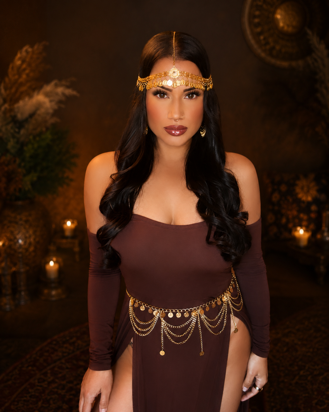 A woman with long black hair wearing gold jewelry, a brown off-shoulder dress, and standing in a dimly lit room with candles and plants.