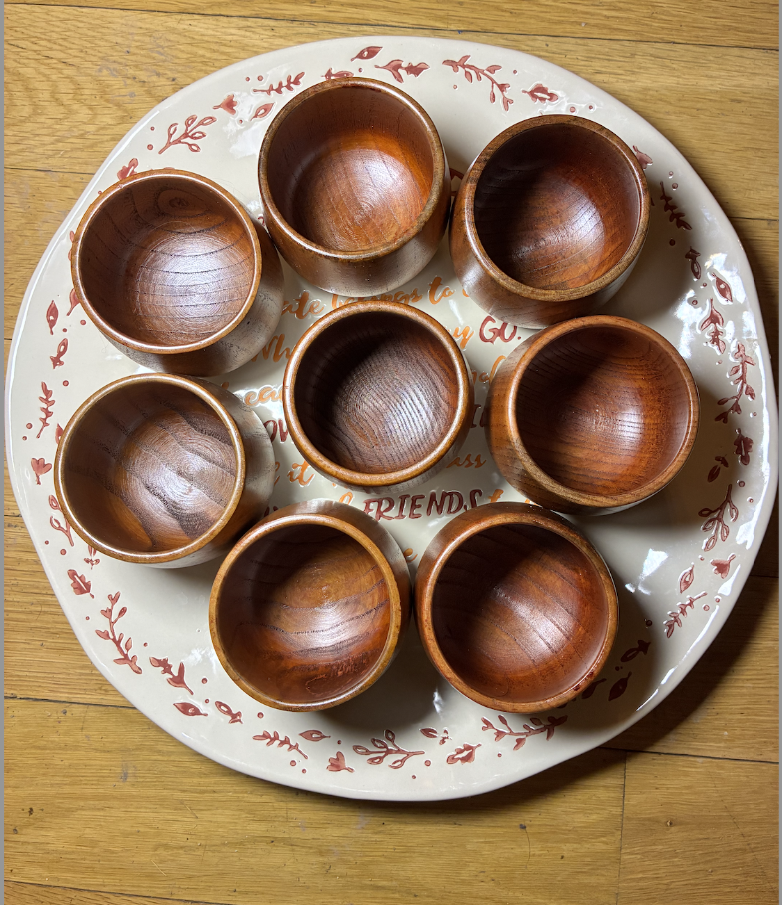 A round white tray with red decorative floral patterns and text underneath, holding nine small wooden bowls on a wooden surface.