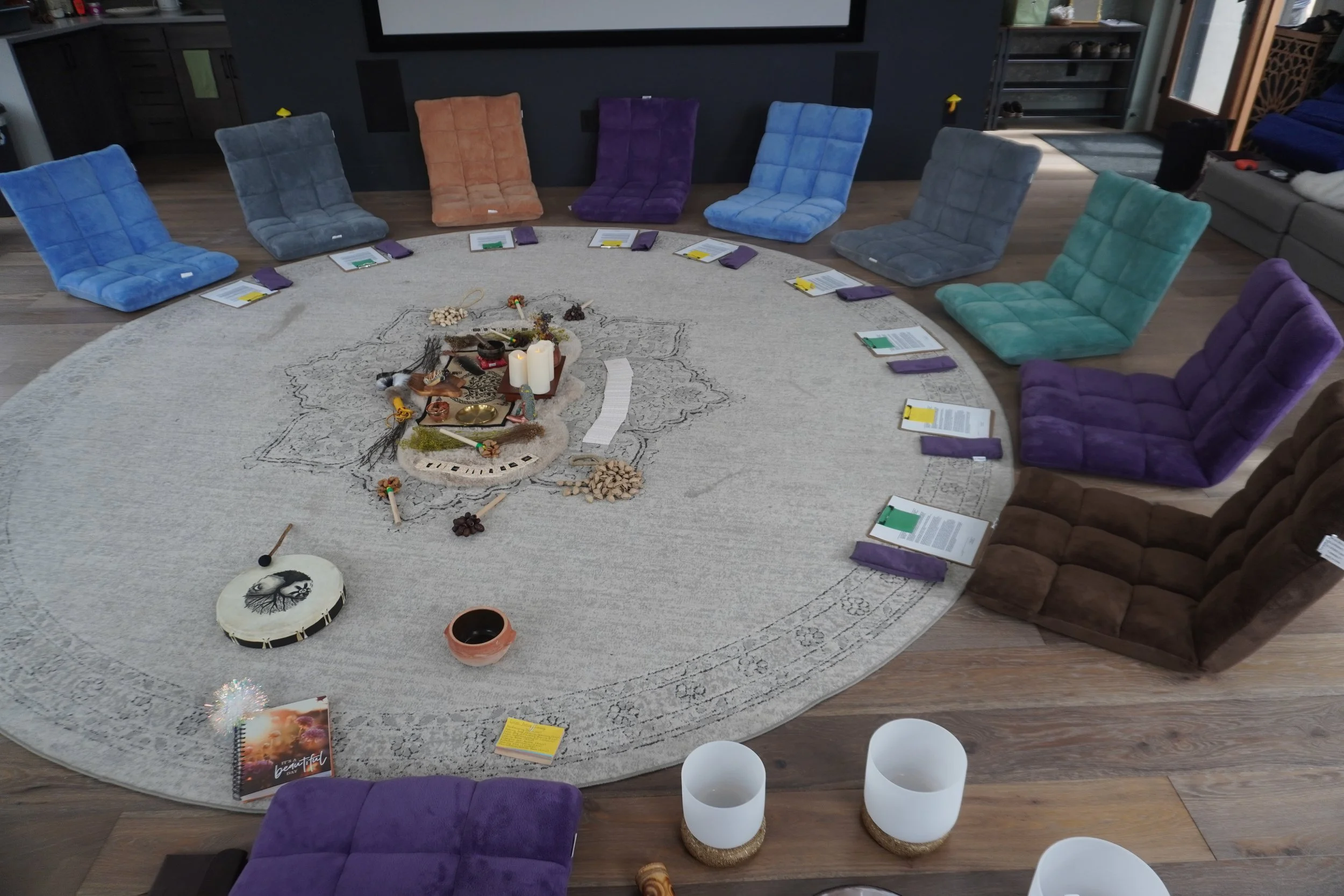 Round living room table with intricately patterned light-colored rug underneath. On the table, there's a centerpiece with candles, crystals, small statues, and decorative items. Seating around the table includes colorful, plush floor chairs in shades