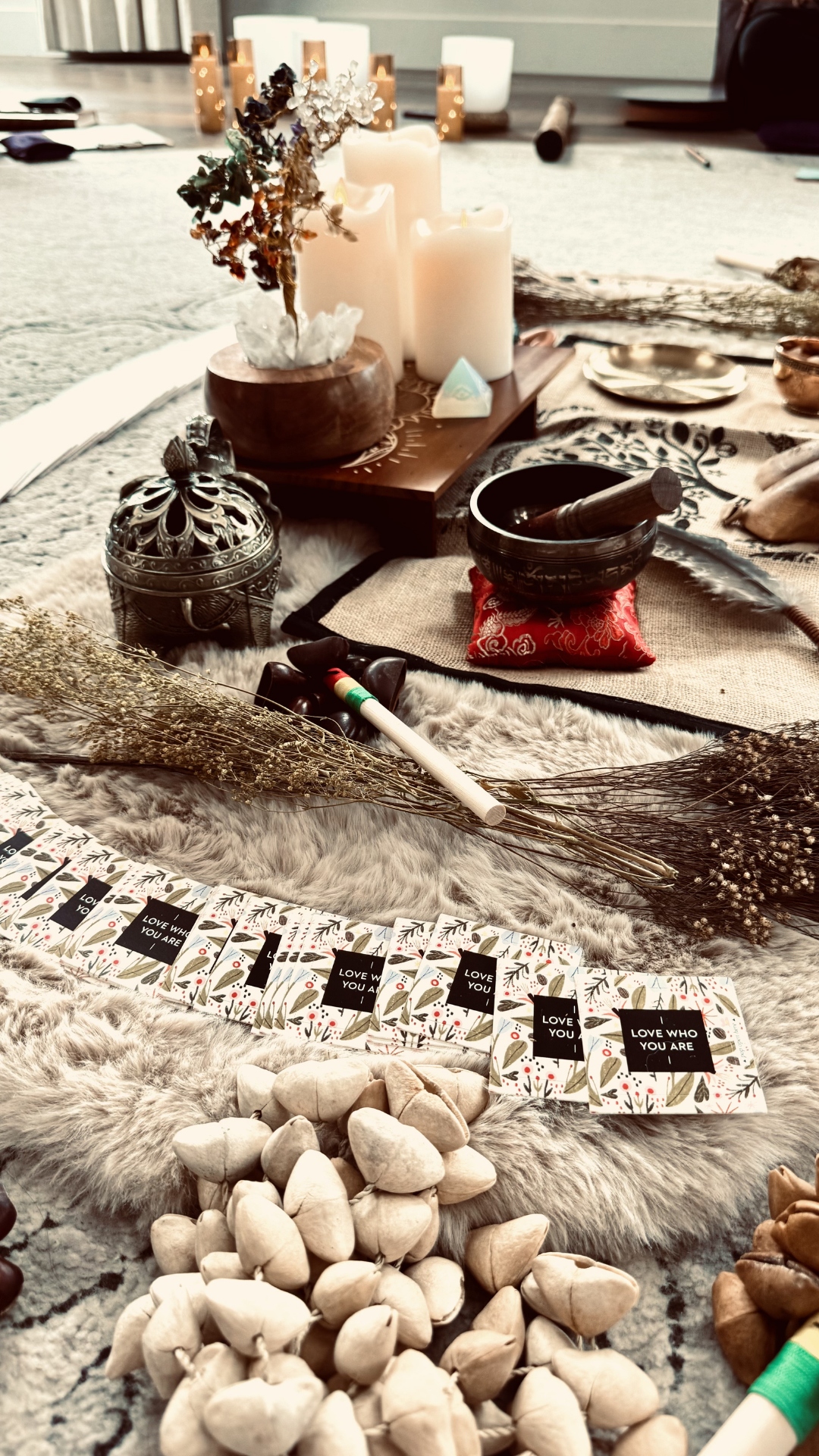 A spiritual or ceremonial altar with candles, dried flowers, a mortar and pestle, stones, and cards with messages like 'Love Who You Are' and 'Love You'.