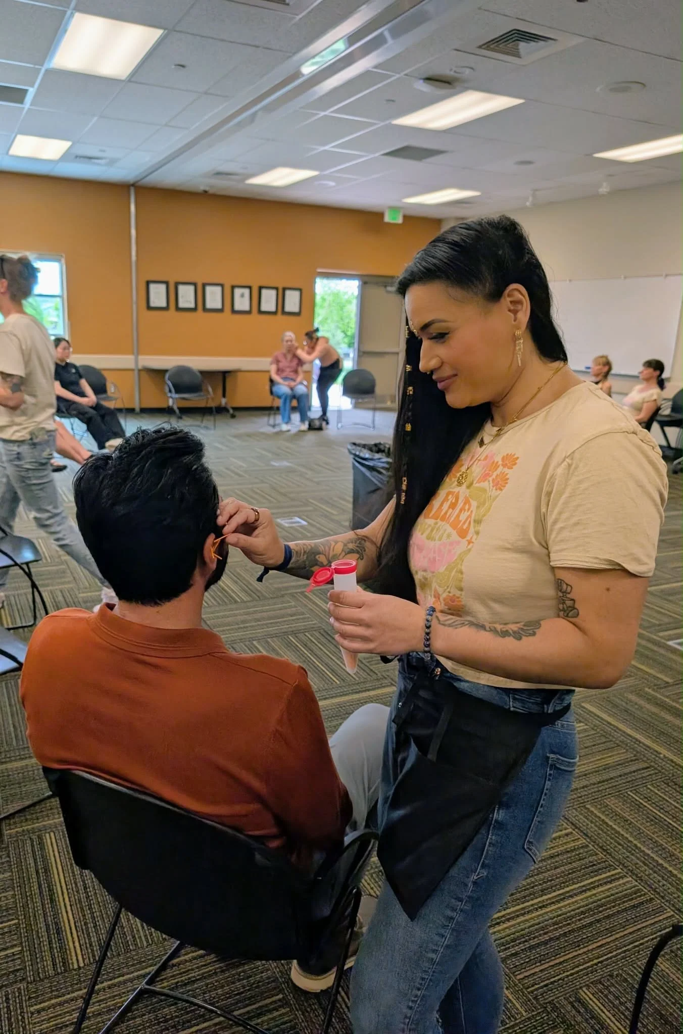 A woman with long black hair, tattoos, and wearing a beige T-shirt with orange print is drawing on the face of a seated person with short dark hair and glasses in a room with several people, chairs, and artwork on the walls.