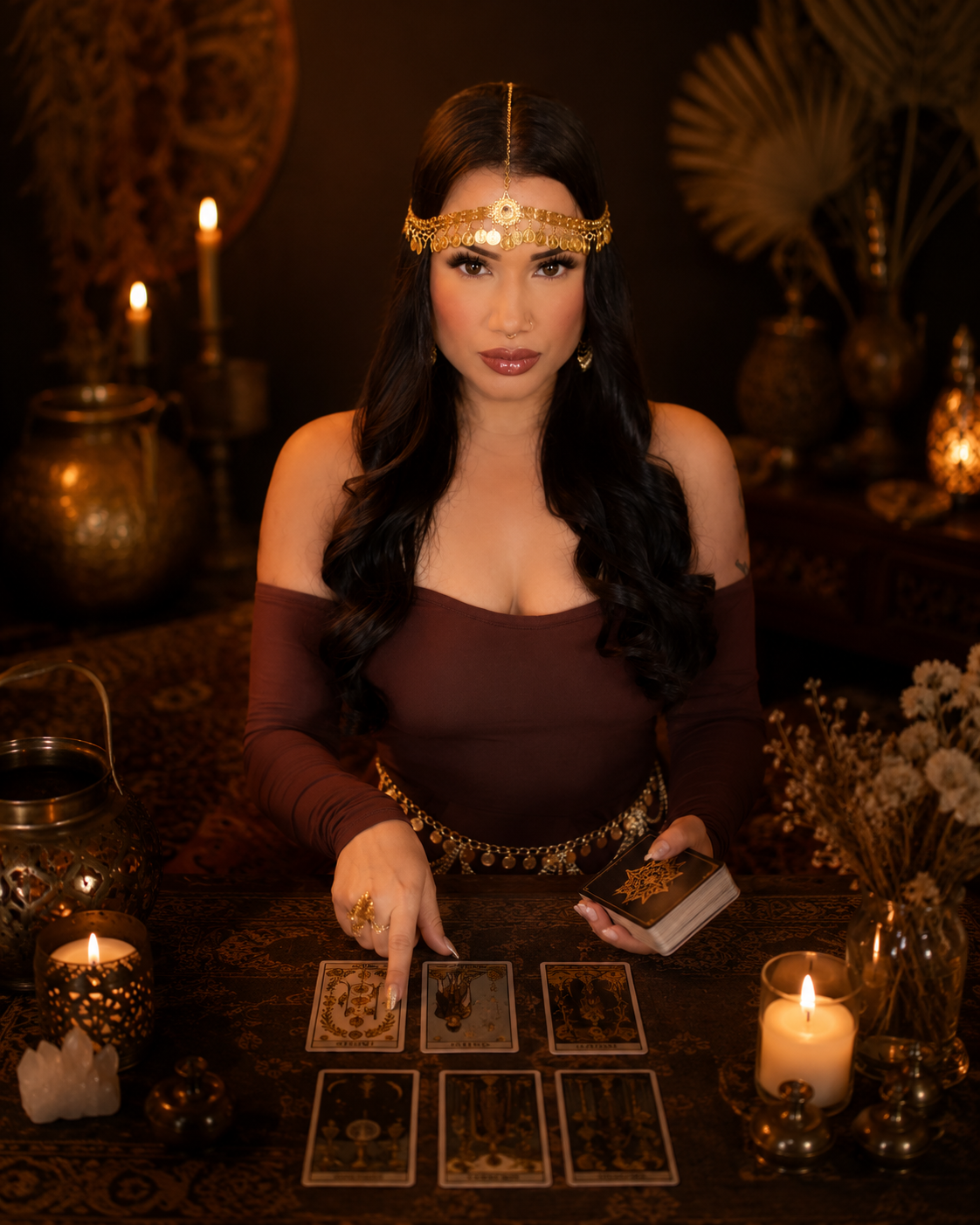 A woman with long dark hair, wearing a gold headpiece and jewelry, sits at a table with tarot cards, surrounded by candles and bohemian decor, performing a tarot reading.