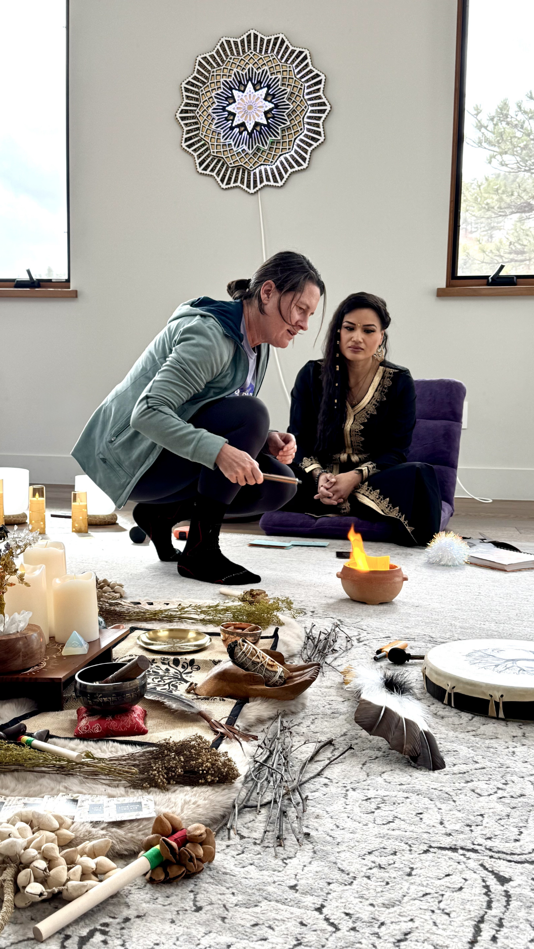 Two women are participating in a spiritual ritual inside a room with two windows, one of which shows trees outside. They are seated on a light-colored carpet surrounded by various objects, including candles, feathers, drums, and dried herbs. One woma