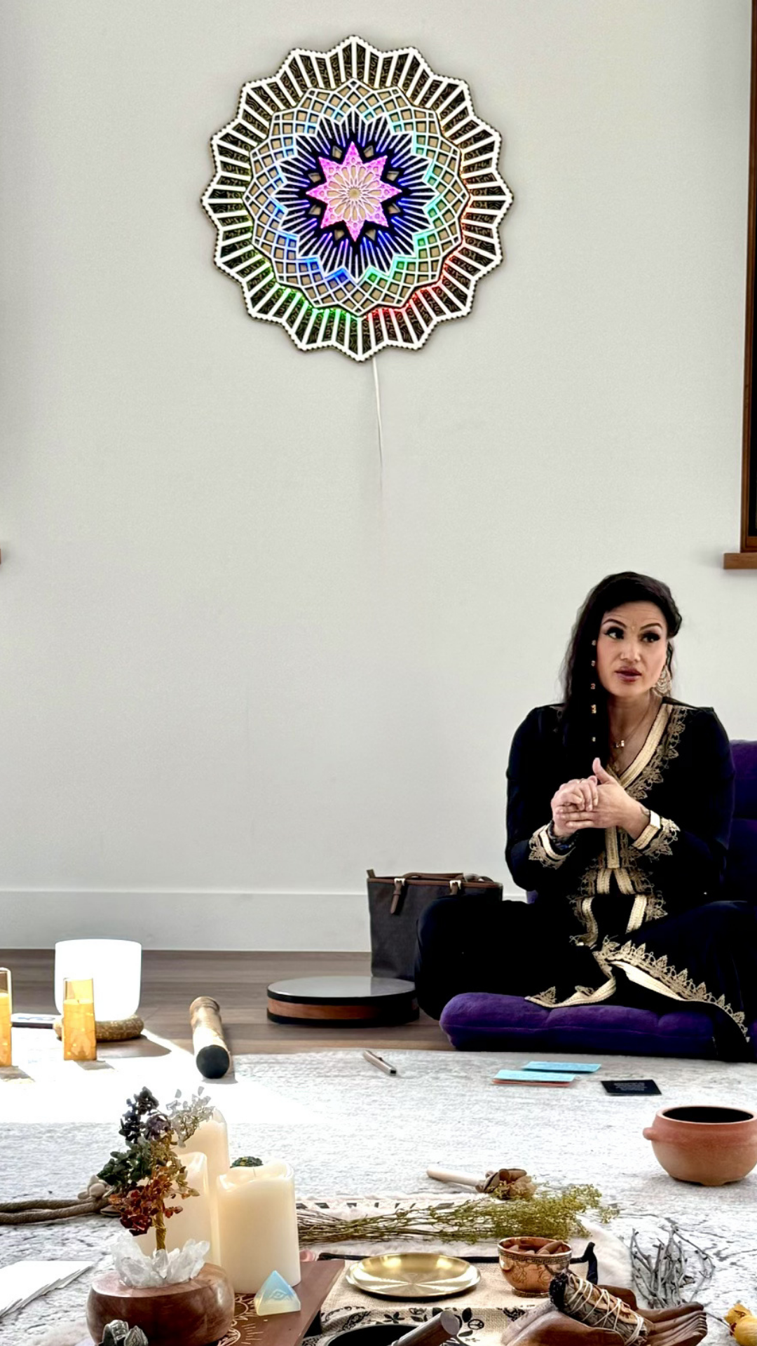 A woman of Asian descent dressed in a black and gold traditional outfit sitting on a purple cushion with her hands clasped. In front of her on a low table are candles, crystals, and decorative items. The background shows a plain wall with colorful ge