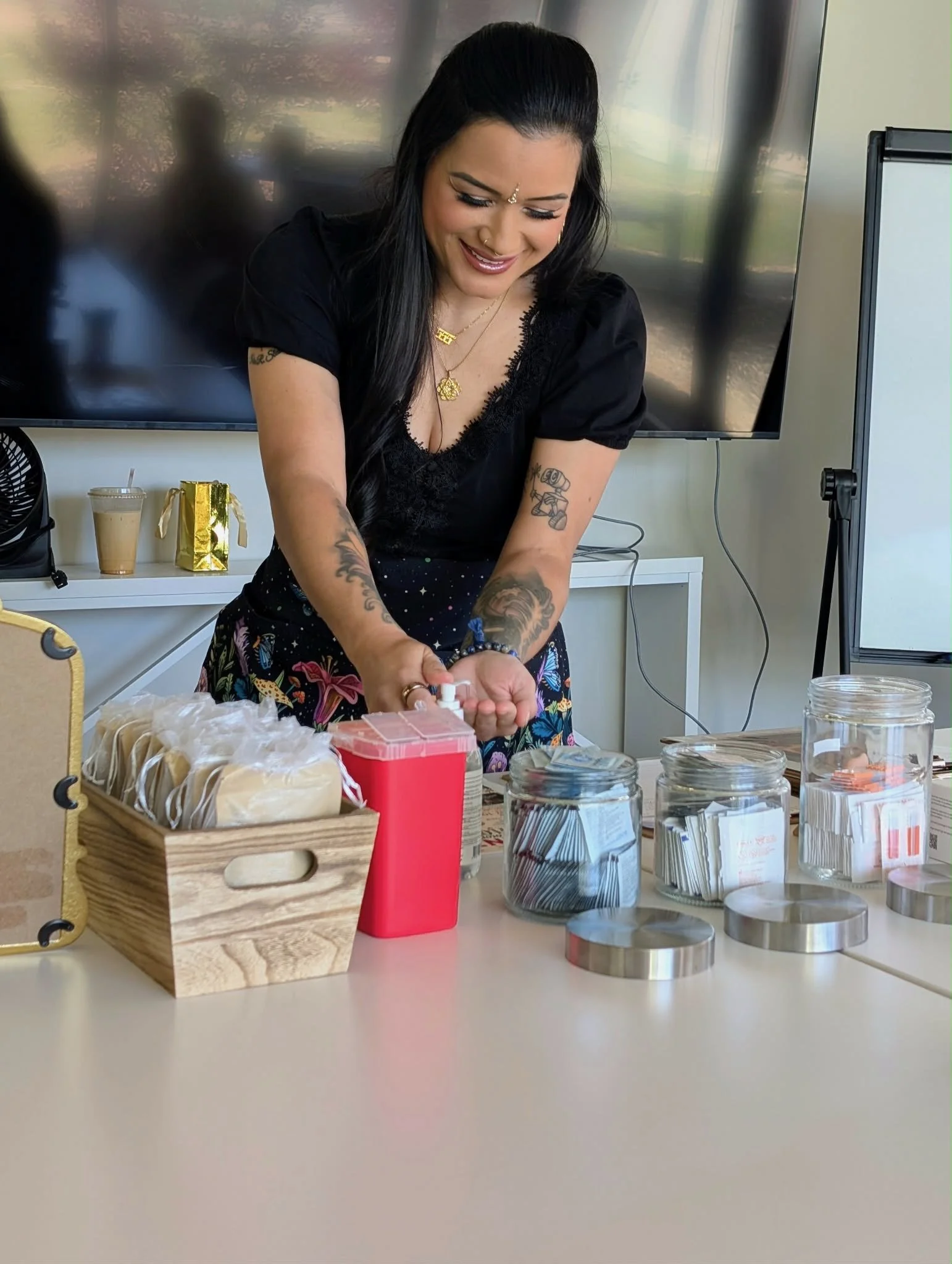 A woman with tattoos on her arms, wearing a black top and floral skirt, is smiling and working at a table filled with various supplies, including jars, tins, a small red box, a wooden crate filled with items, and a white paper bag, in a room with a b