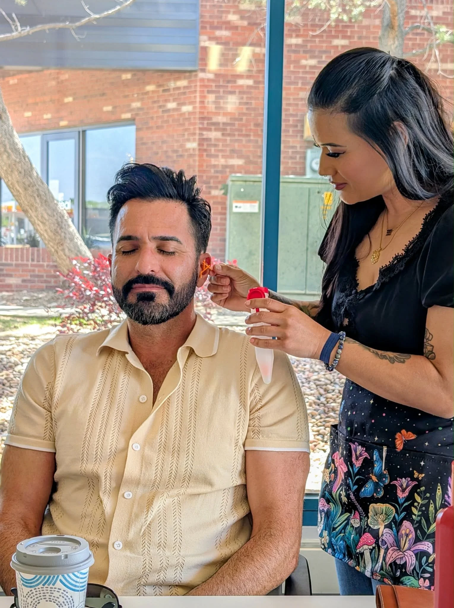 A woman is placing a hearing aid in a man's ear at an outdoor setting with a brick building and trees in the background.