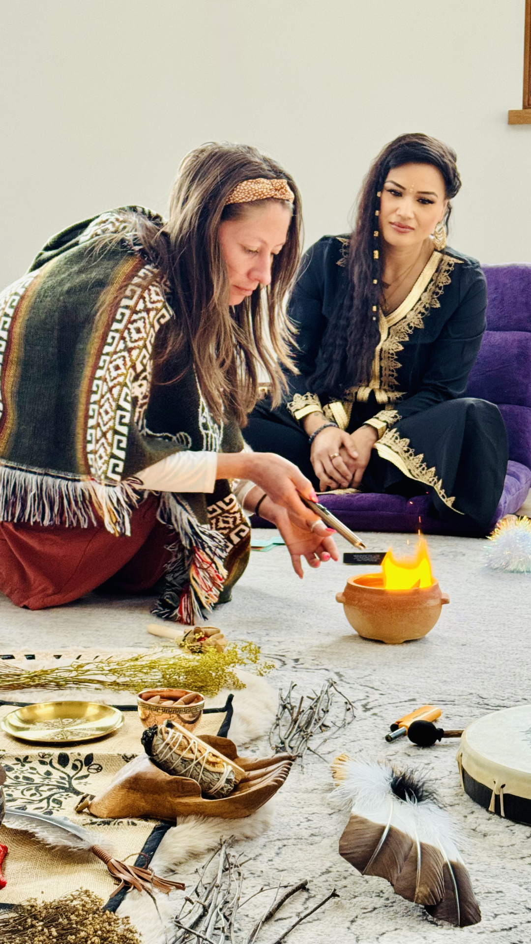 Two women engaging in a spiritual or ceremonial ritual indoors. One woman is dressed in traditional or colorful attire and is holding a small object over a clay bowl with a flame. The other woman dressed in black with gold embroidery is sitting nearb