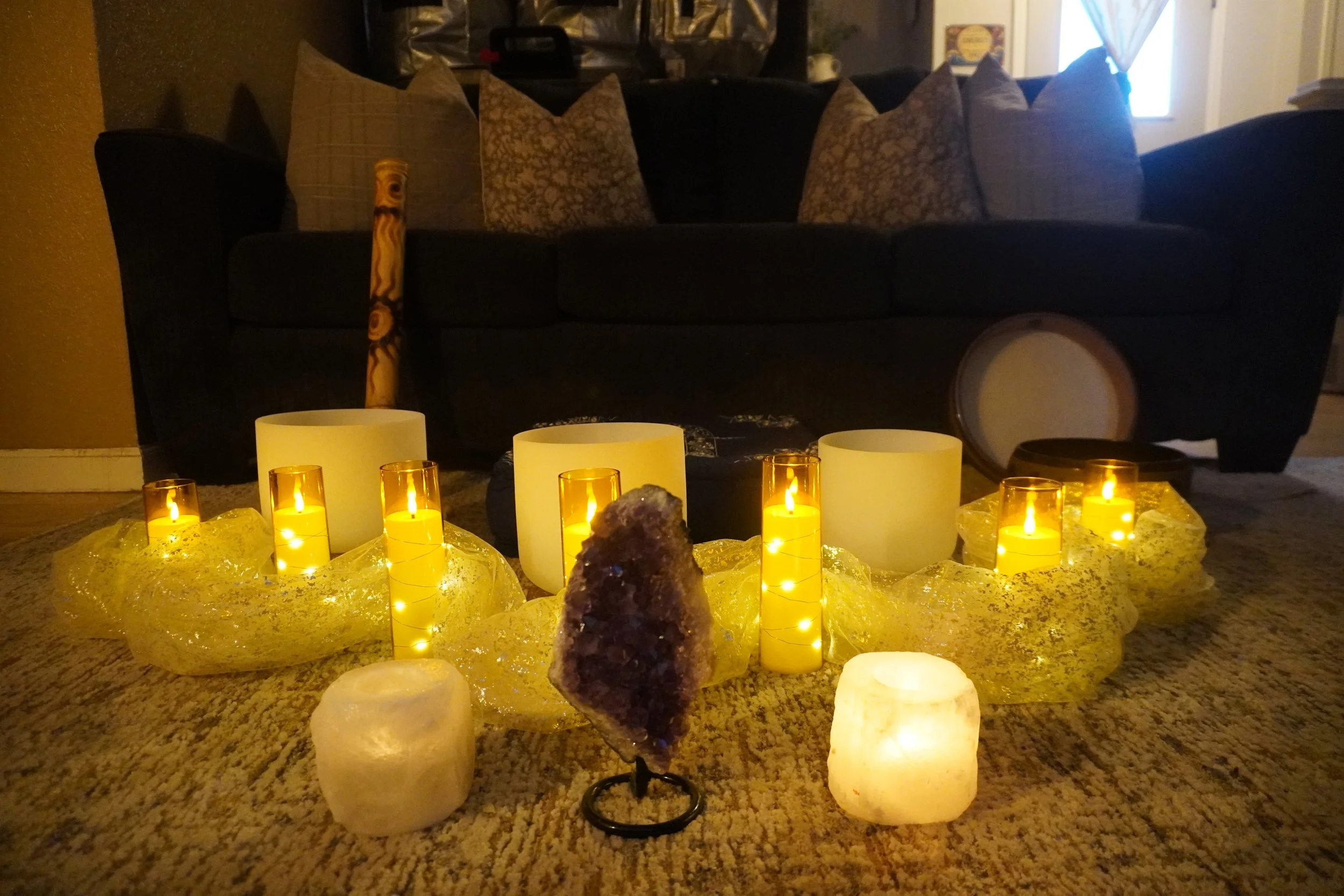 A living room decorated for a cozy, warm atmosphere with numerous candles on the floor emitting a yellow glow, with pillows on a dark-colored sofa in the background, and a window with curtains letting in some light.