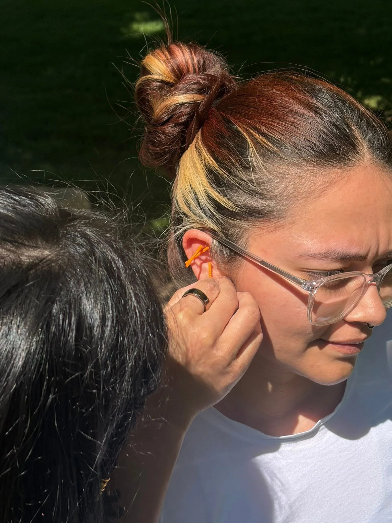 Close-up of a woman with glasses having her ear pierced by a person with dark hair.