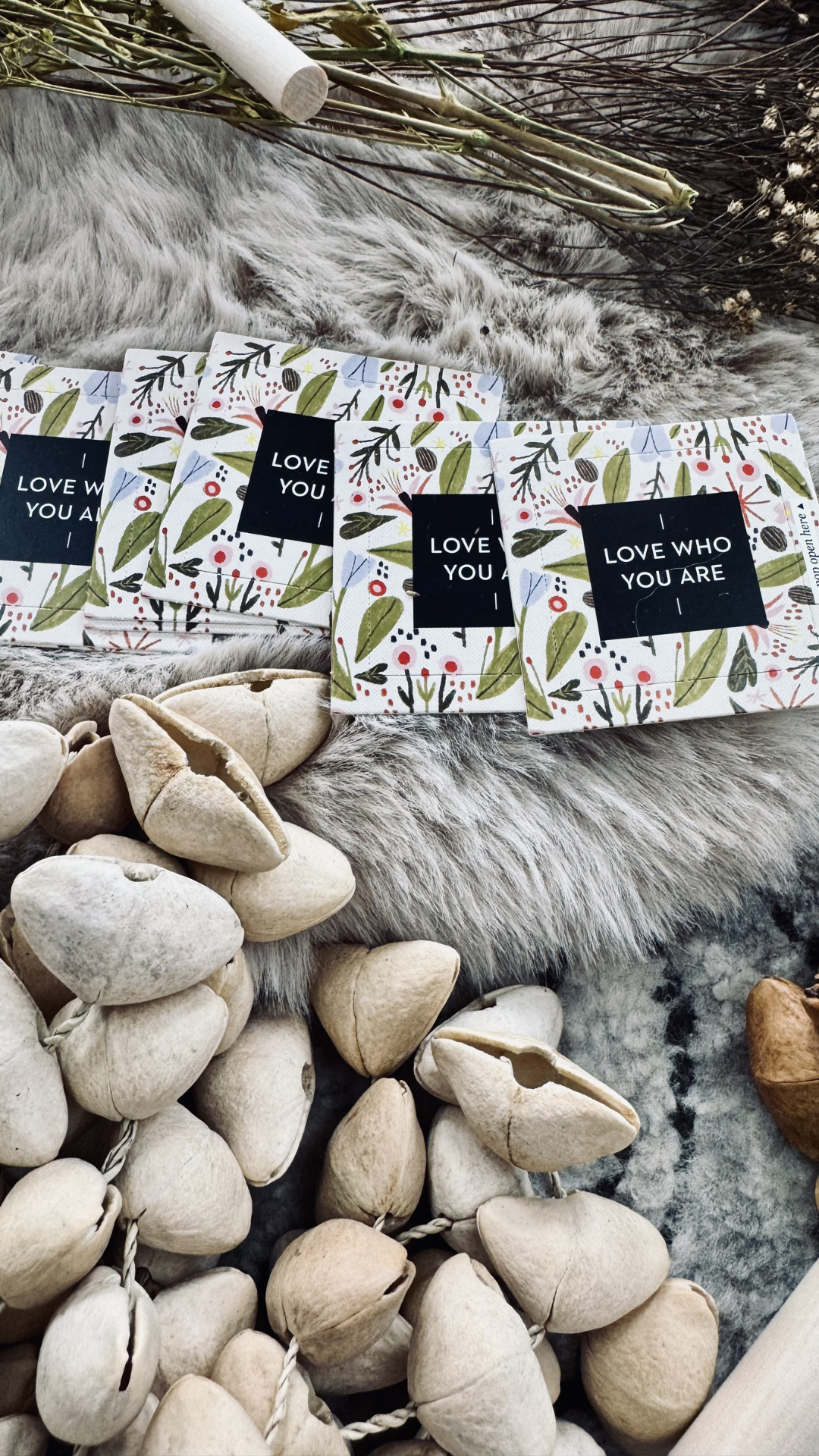 Valentine's Day or romantic-themed greeting cards with floral designs and black squares with white text, placed on a faux fur surface with dried plants and shells nearby.