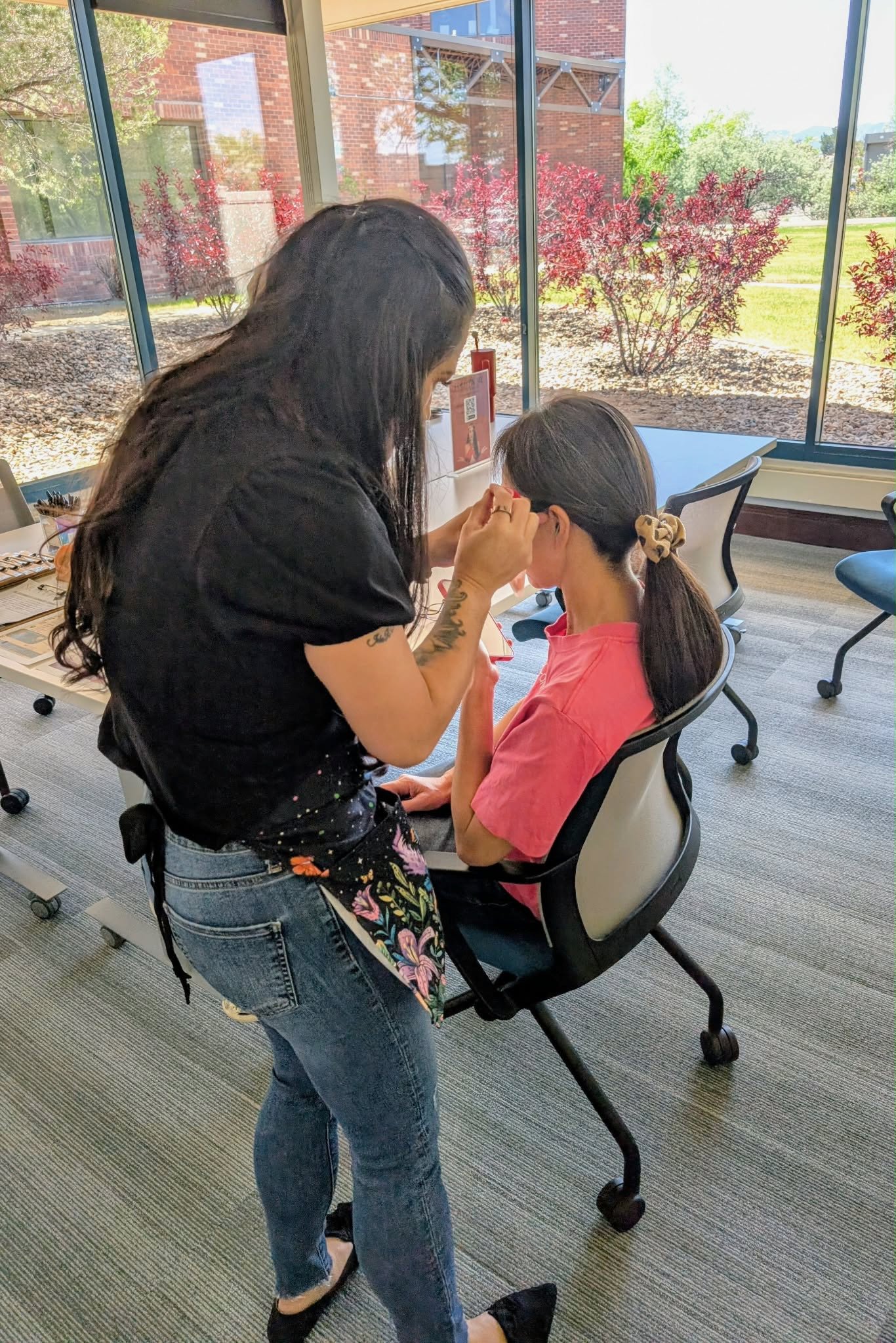A woman with long dark hair and tattoos on her arm is standing, leaning over, and helping another woman, who is seated and has long dark hair tied back with a large scrunchie, while she looks at her phone. They are inside a room with large windows re