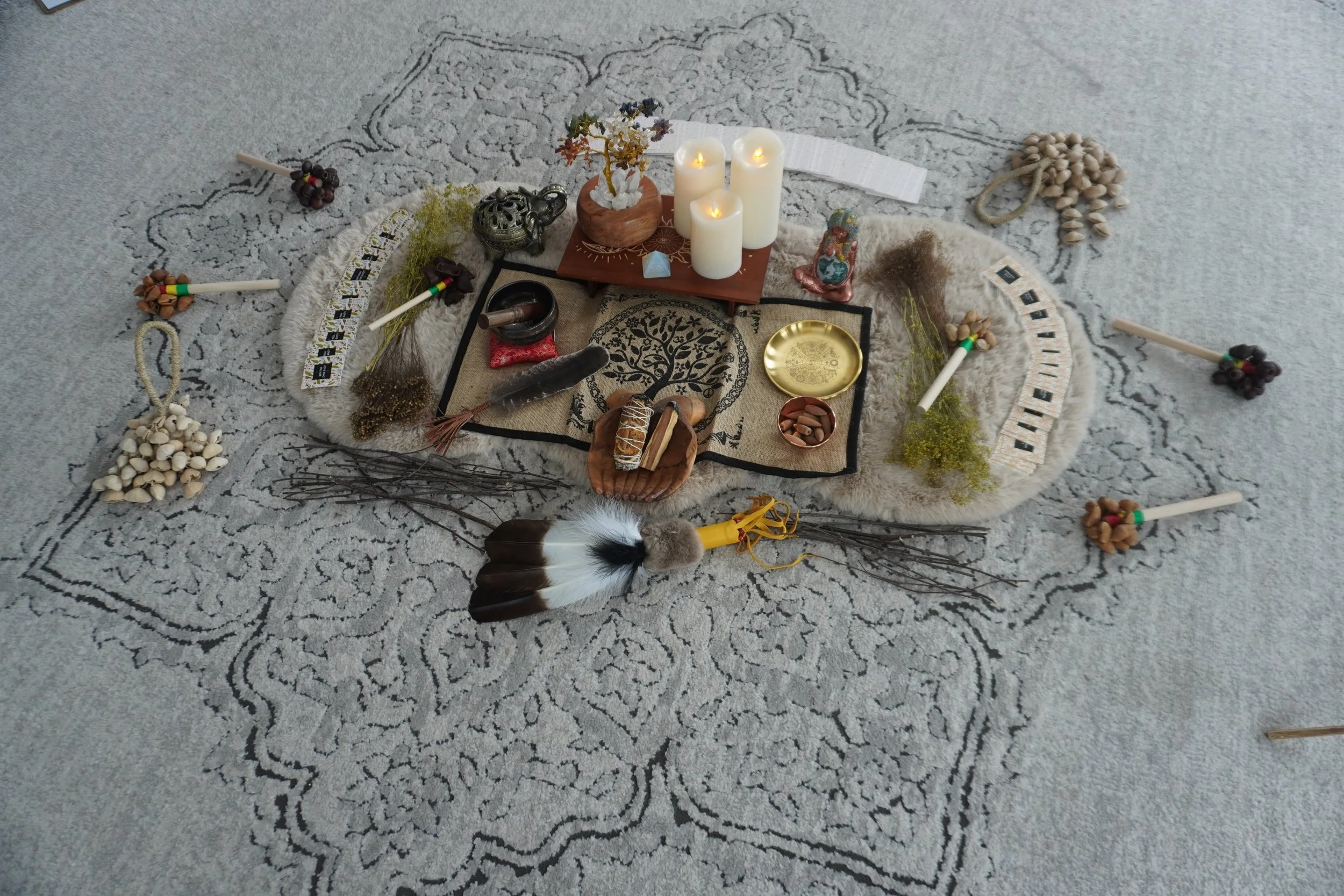 A small altar setup on a patterned rug with white candles, a small plant, decorative objects, and various spiritual tools, surrounded by bundles of dried herbs and small stone-like objects.