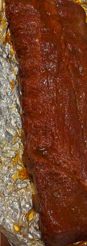 A rack of pork ribs covered in barbecue sauce on aluminum foil.