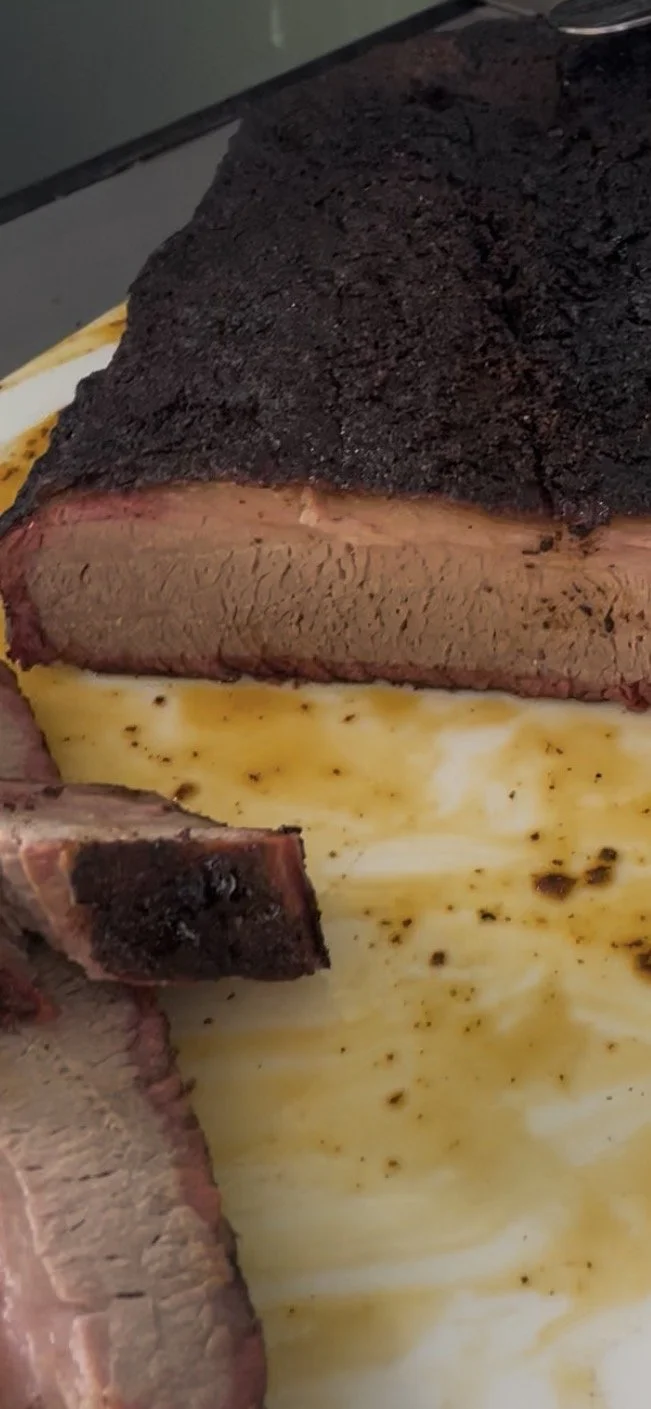 Close-up of a sliced beef brisket with a dark bark crust, served on a white plate with brown sauce drizzled on it.