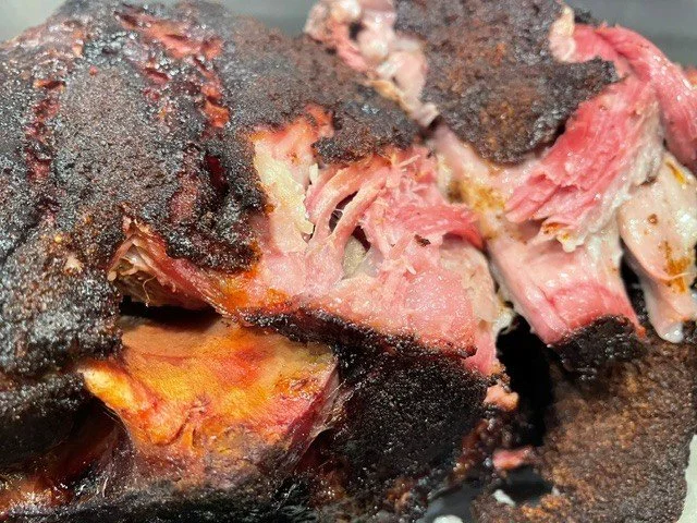 Close-up of a smoked or roasted piece of meat with a charred crust, partially sliced to reveal pink, tender interior.