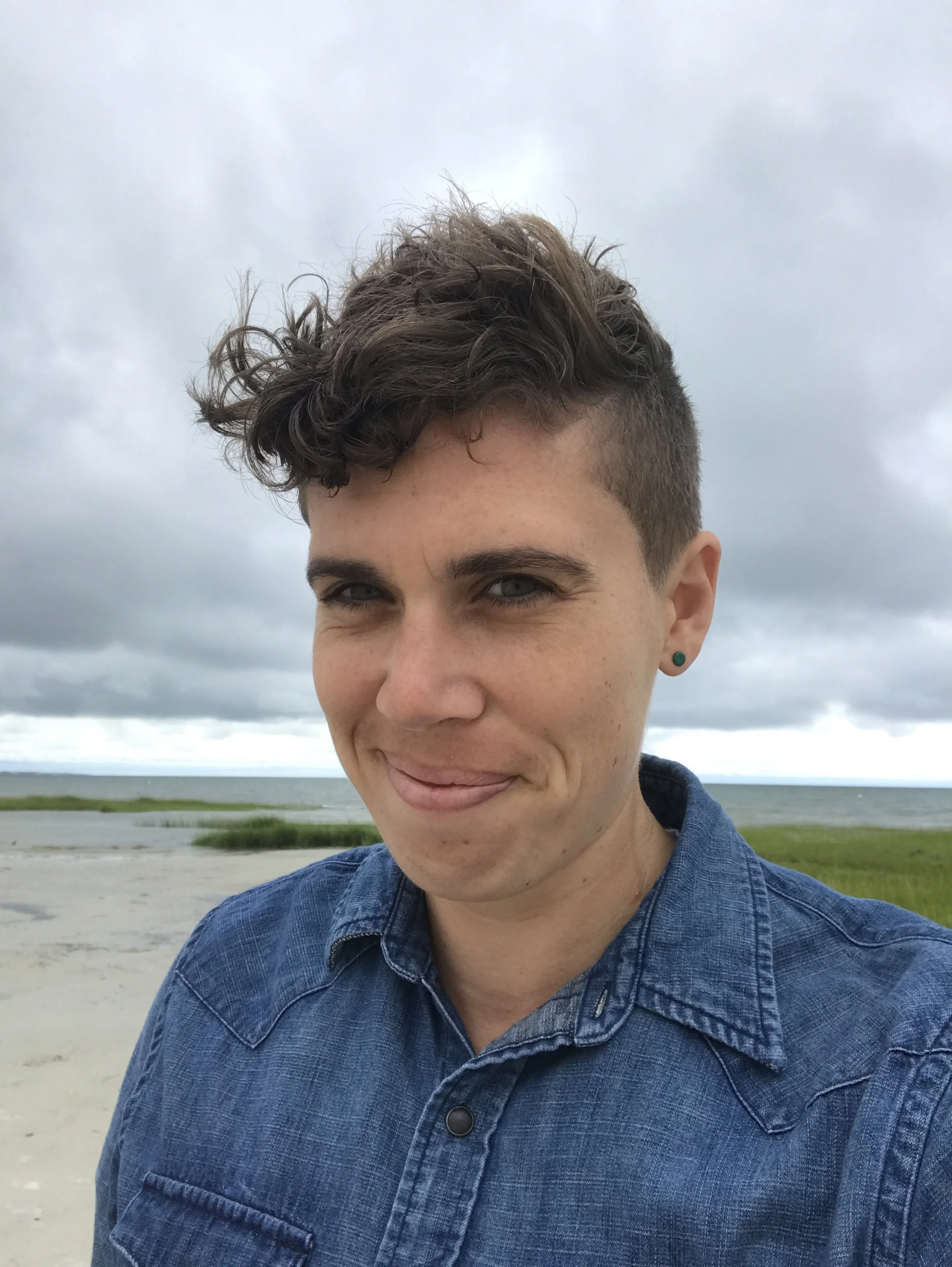 Close-up of a person with short, curly hair and piercing blue eyes, wearing a denim jacket, outdoors on a cloudy day with water and grassy land in the background.