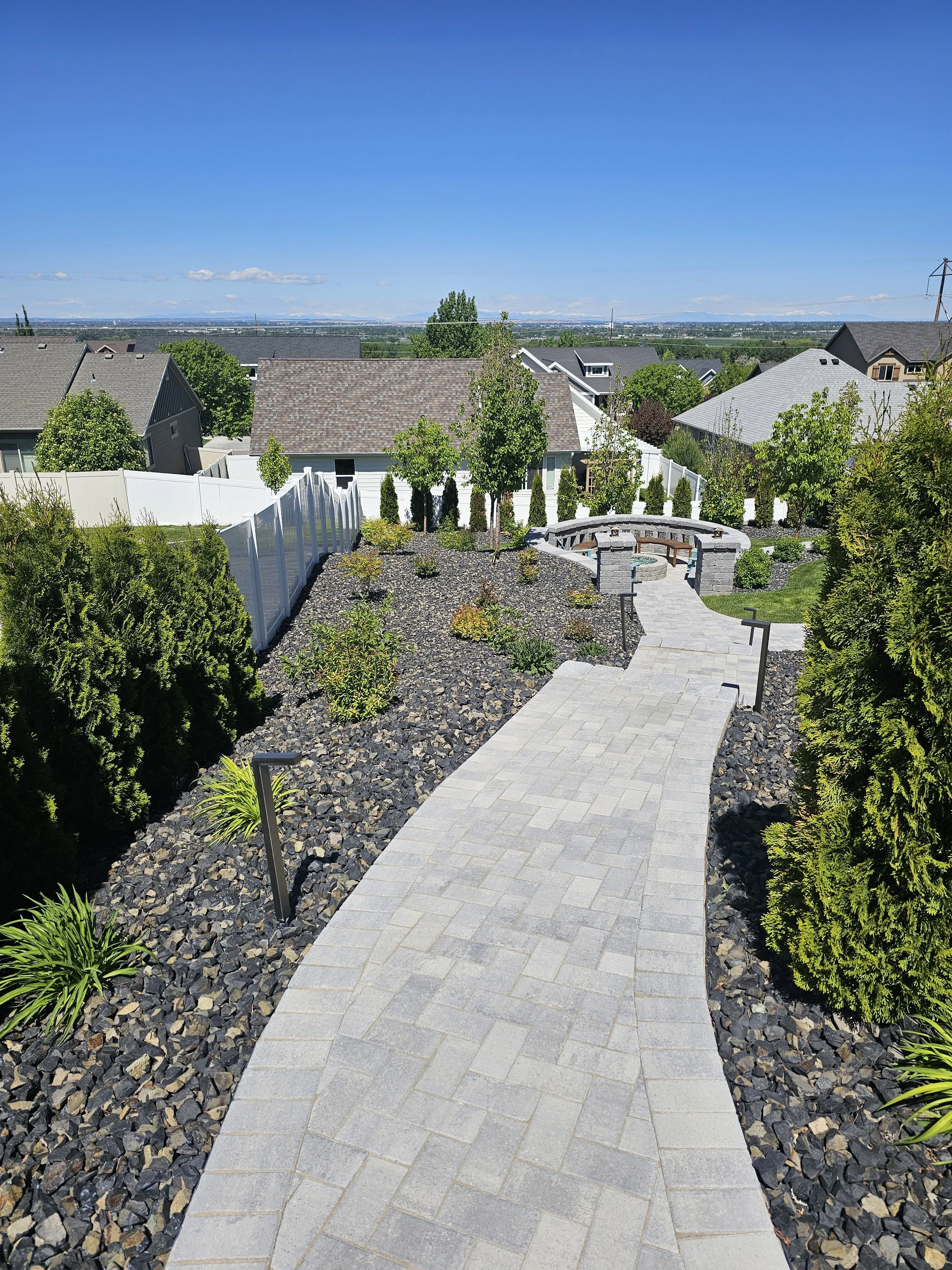 A backyard with a paved walkway, landscaped with bushes and trees, enclosed by a white fence. There's a stone fire pit with benches to the right, and neighboring houses with gray and brown roofs under a clear blue sky.
