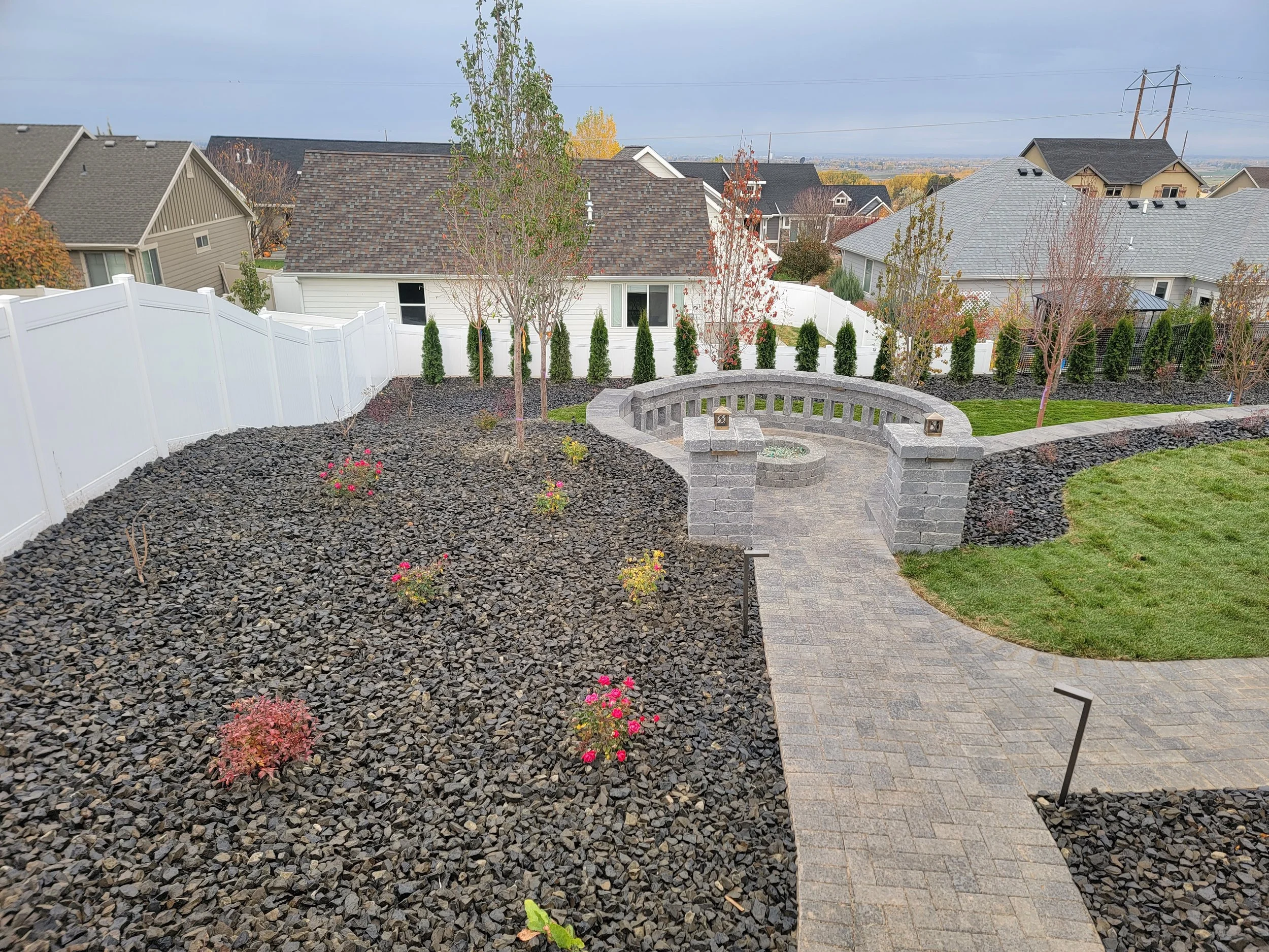 A backyard garden with a curved stone bench, small trees, and flower bushes, enclosed by a white fence, with neighboring houses and an overcast sky in the background.