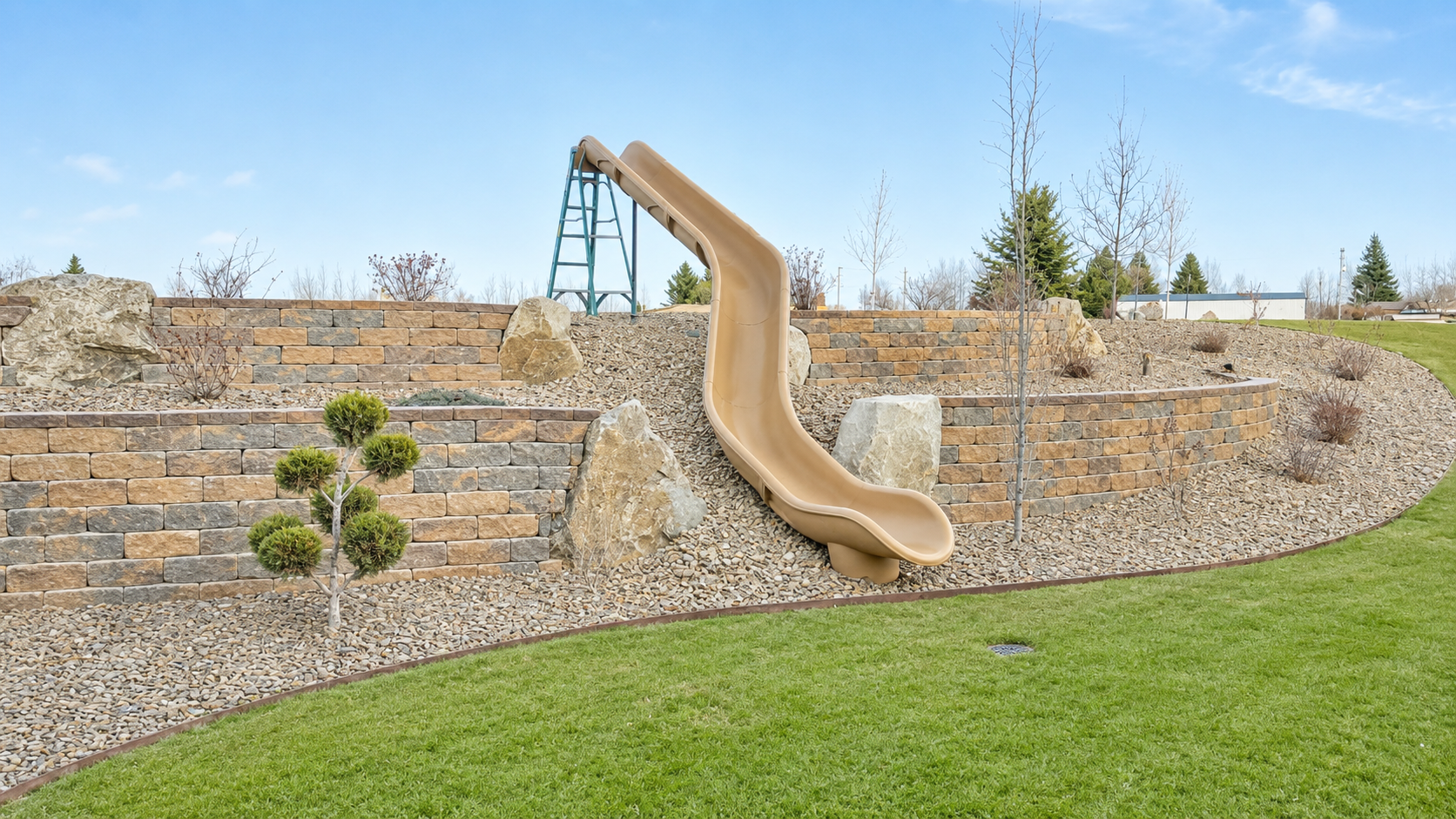 A backyard area with a tan slide on a gravel and stone landscaped hill, steps made of brick, some small trees and shrubs, green grass in the foreground, and a blue sky with clouds.