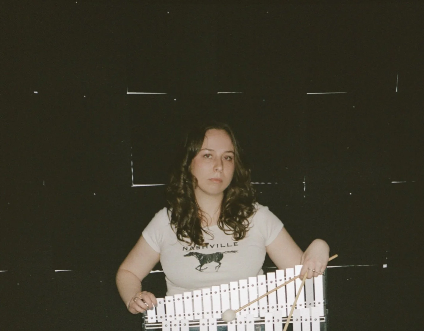 A woman with curly brown hair playing a white xylophone against a dark background with faint light markings.