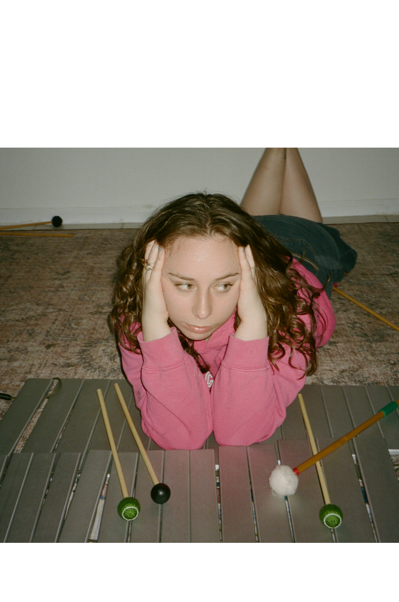 A young woman with curly brown hair lying on her stomach on the floor, her head supported by her hands, with a xylophone in front of her and colorful mallets scattered around.