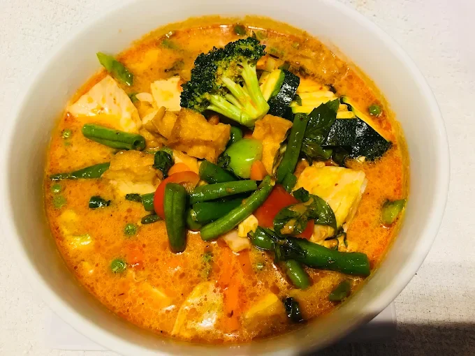 A bowl of vegetable curry with broccoli, zucchini, green beans, carrots, and tofu in a spicy, orange-colored sauce.