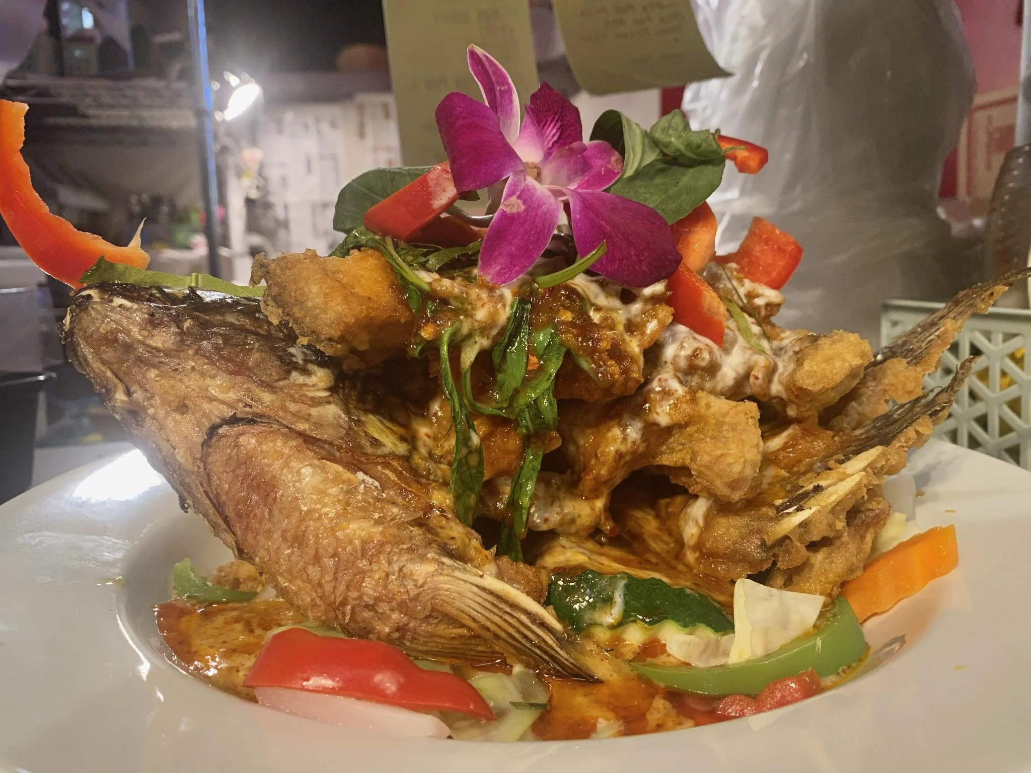 A plate of fried fish topped with a colorful salad that includes pink edible flowers, lettuce, sliced red peppers, and other vegetables, with a sauce or dressing near the fish.