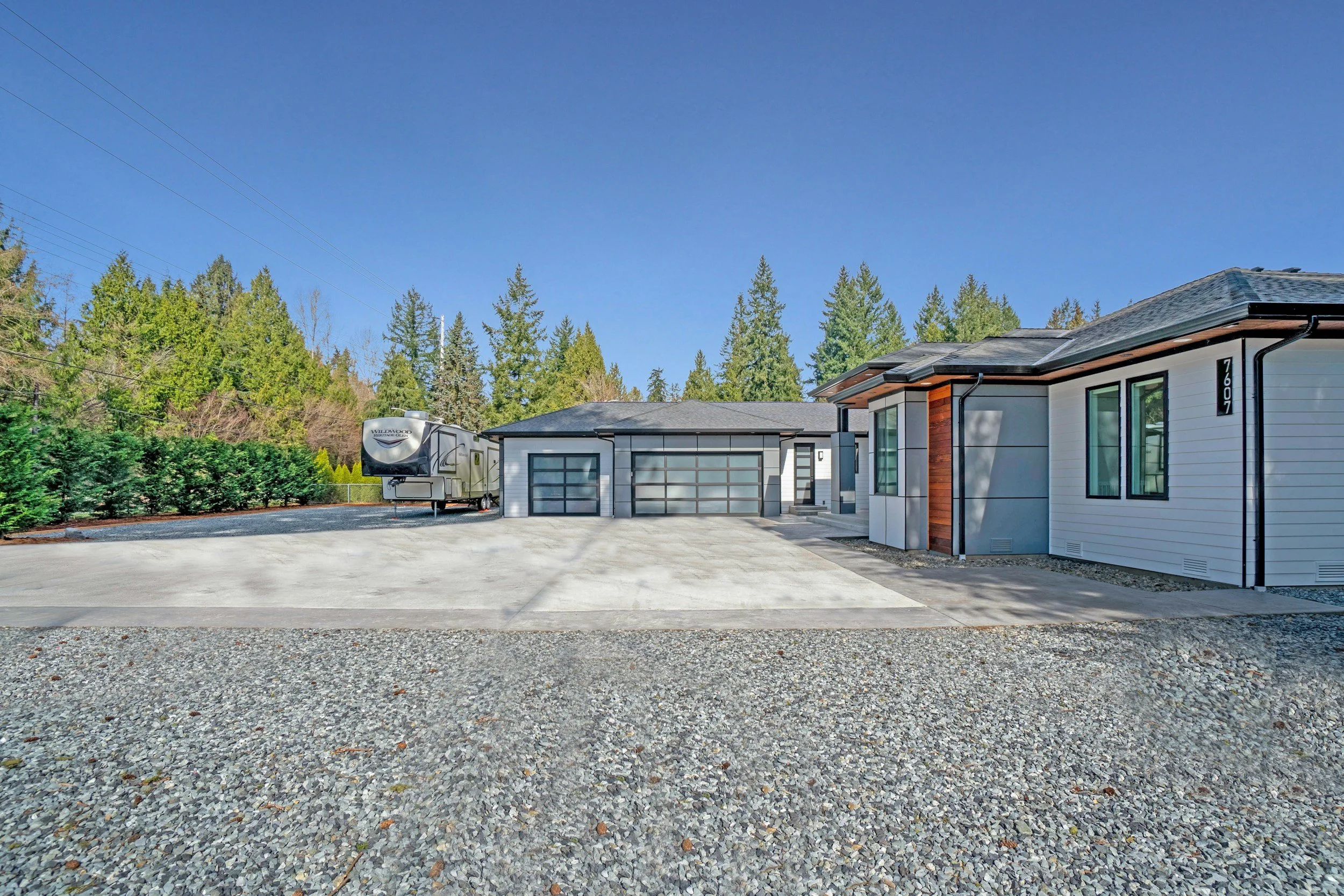 Modern house with a gray garage door, driveway, and adjacent white building with large windows, surrounded by trees and a clear blue sky.