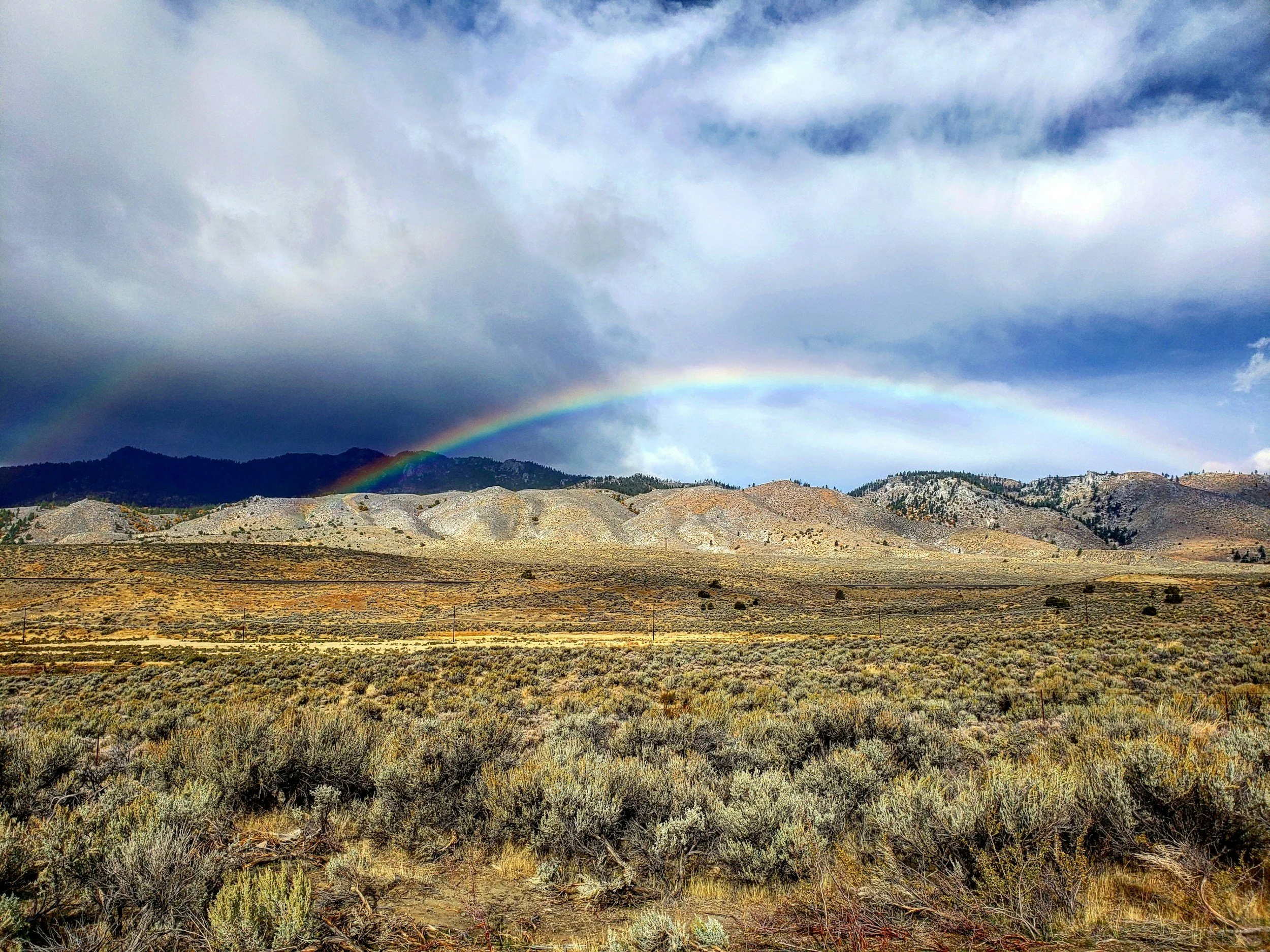 Scenic view of a mountainous landscape with a rainbow in the sky and clouds overhead. The foreground shows dry shrubs and a fence line.