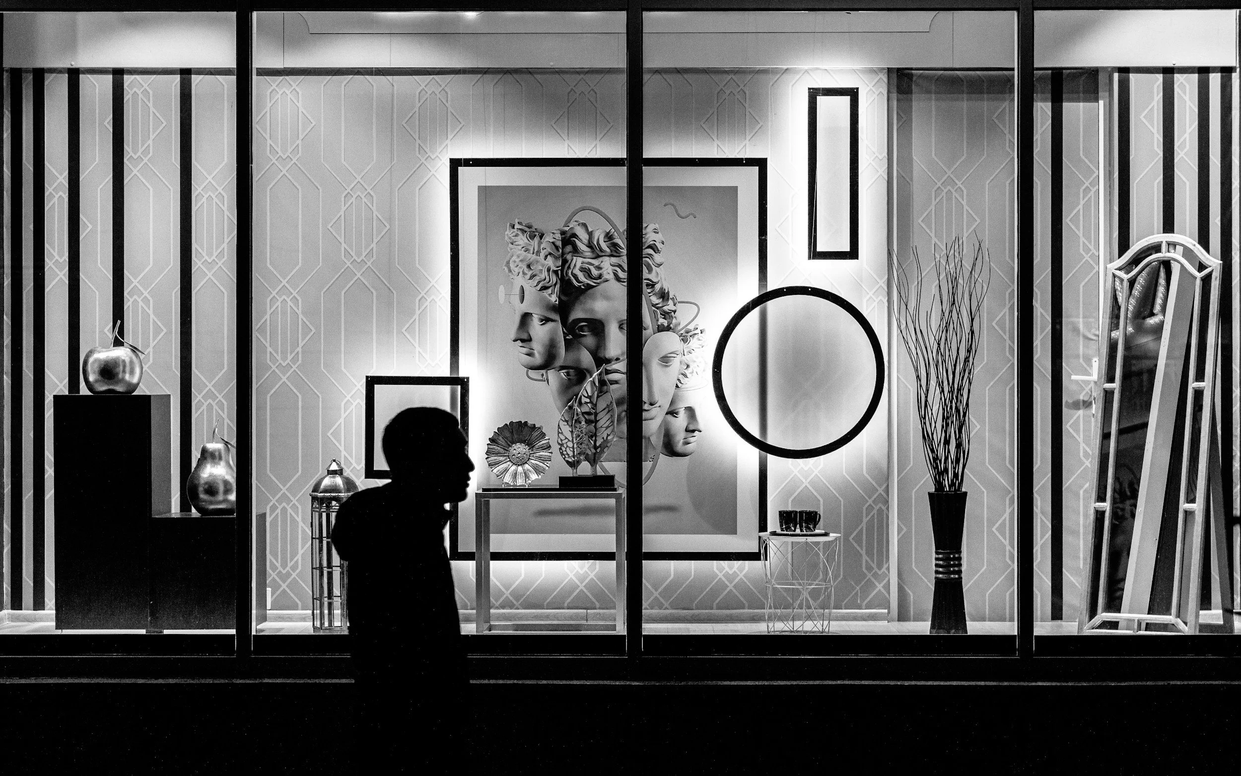 A black and white photo of an art gallery window display featuring sculptures and framed artwork, with a silhouette of a person in front.