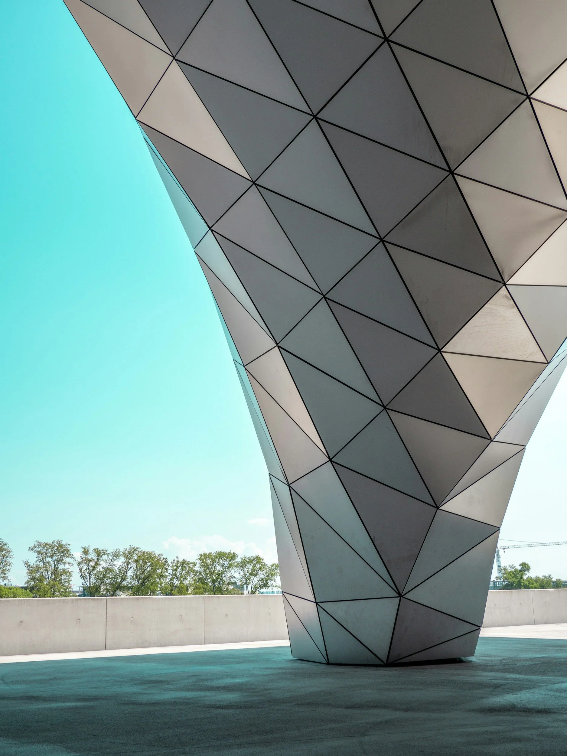 A modern architectural structure with a geometric, triangular pattern on its exterior, set against a clear blue sky and distant trees.