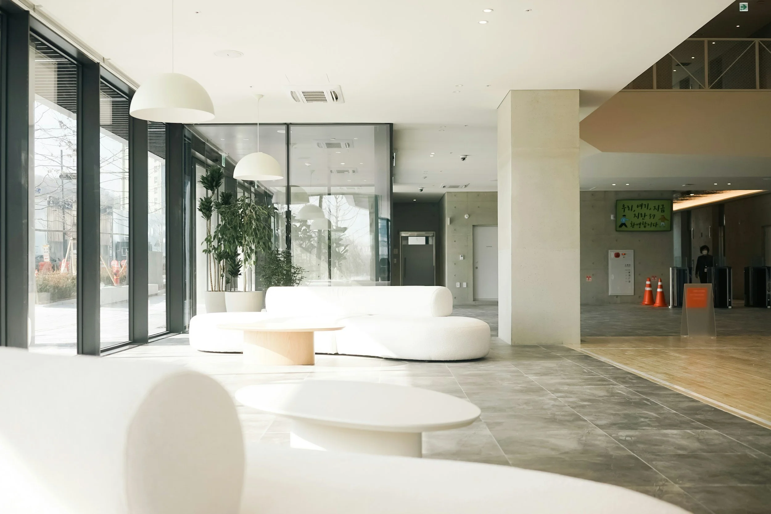 Modern indoor lobby with white seating, large windows, and indoor plants, illuminated by natural light and hanging pendant lights.